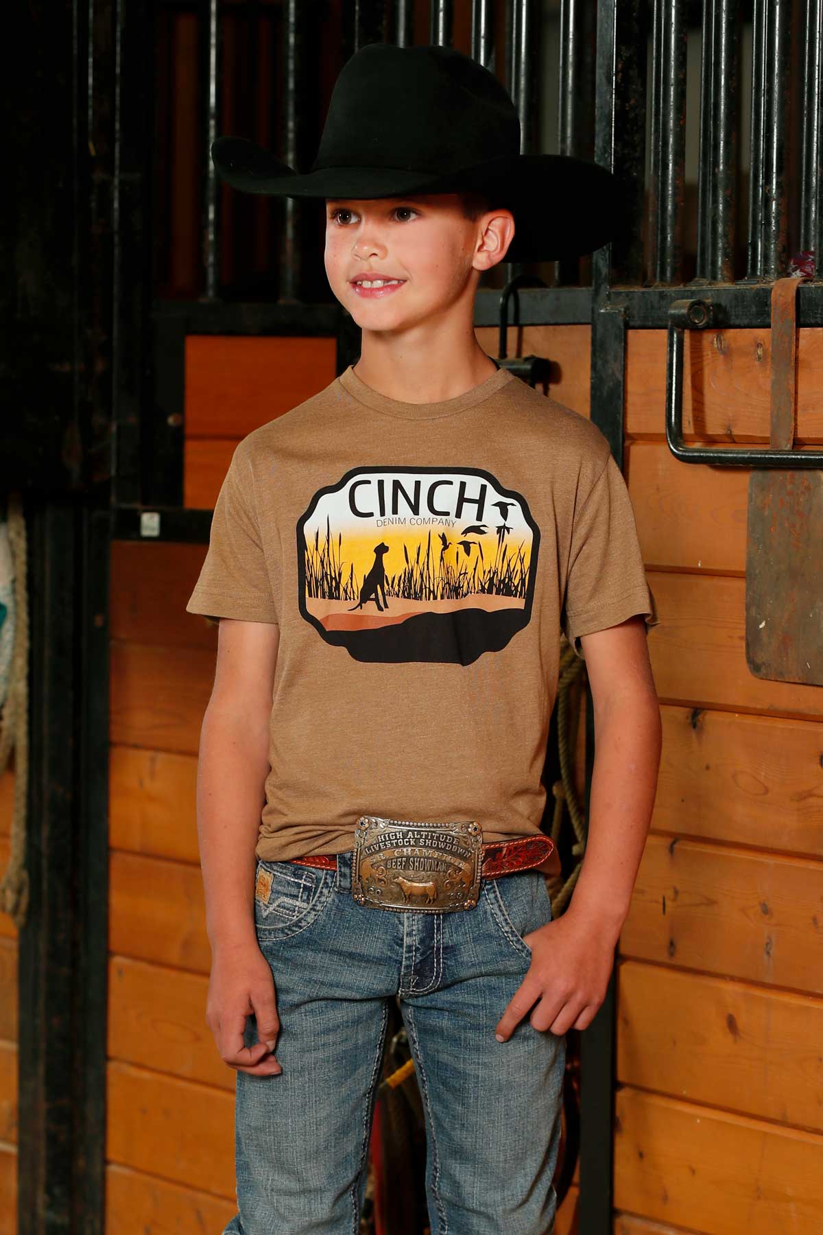 Young boy standing in a barn wearing a 'CINCH' t-shirt with a western graphic, paired with jeans and a cowboy hat