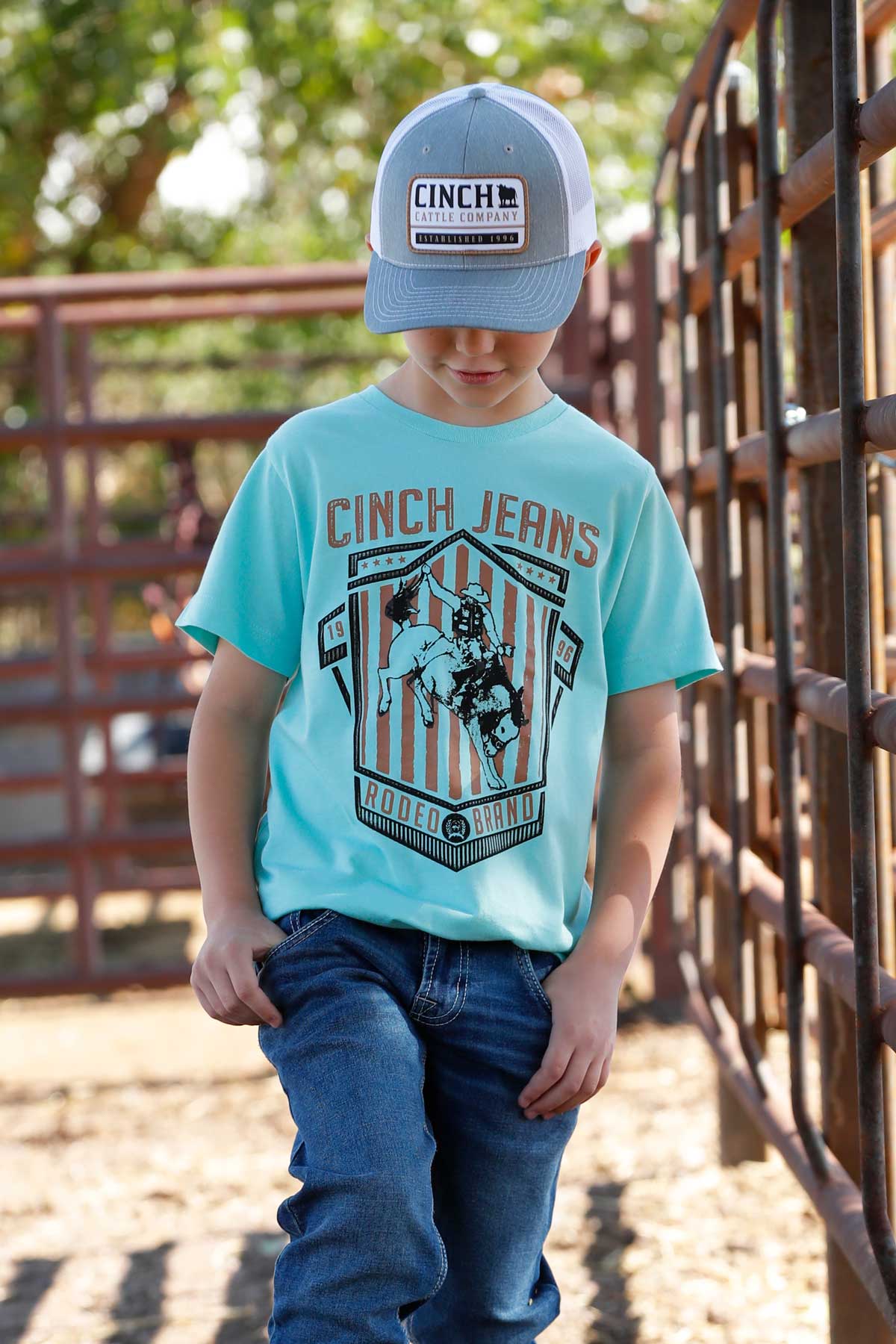 Young boy standing outside wearing a teal shirt with a western graphic, with jeans and a hat