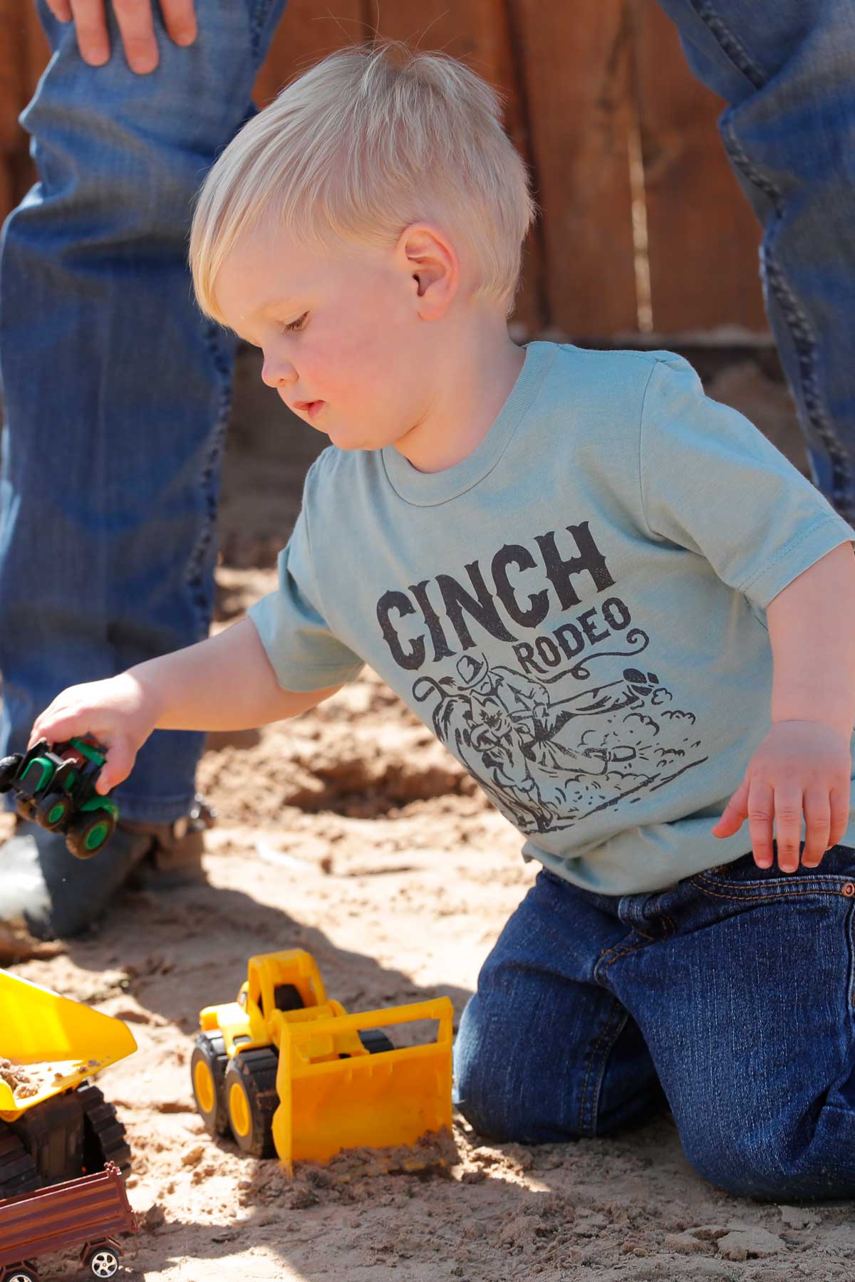 Little boy playing with toy trucks outside, wearing a green 'CINCH' t-shirt and jeans