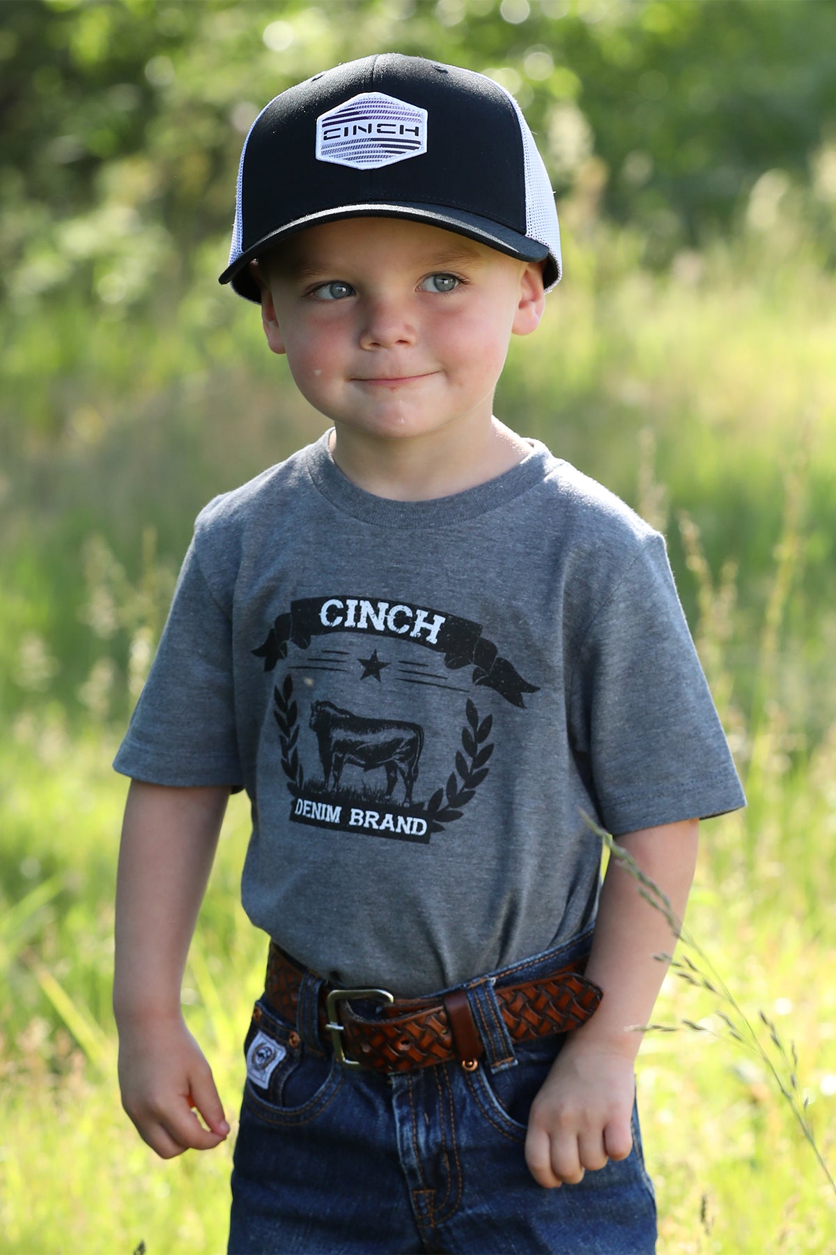 Little boy standing outside wearing a gray t-shirt with jeans, a belt, and a hat