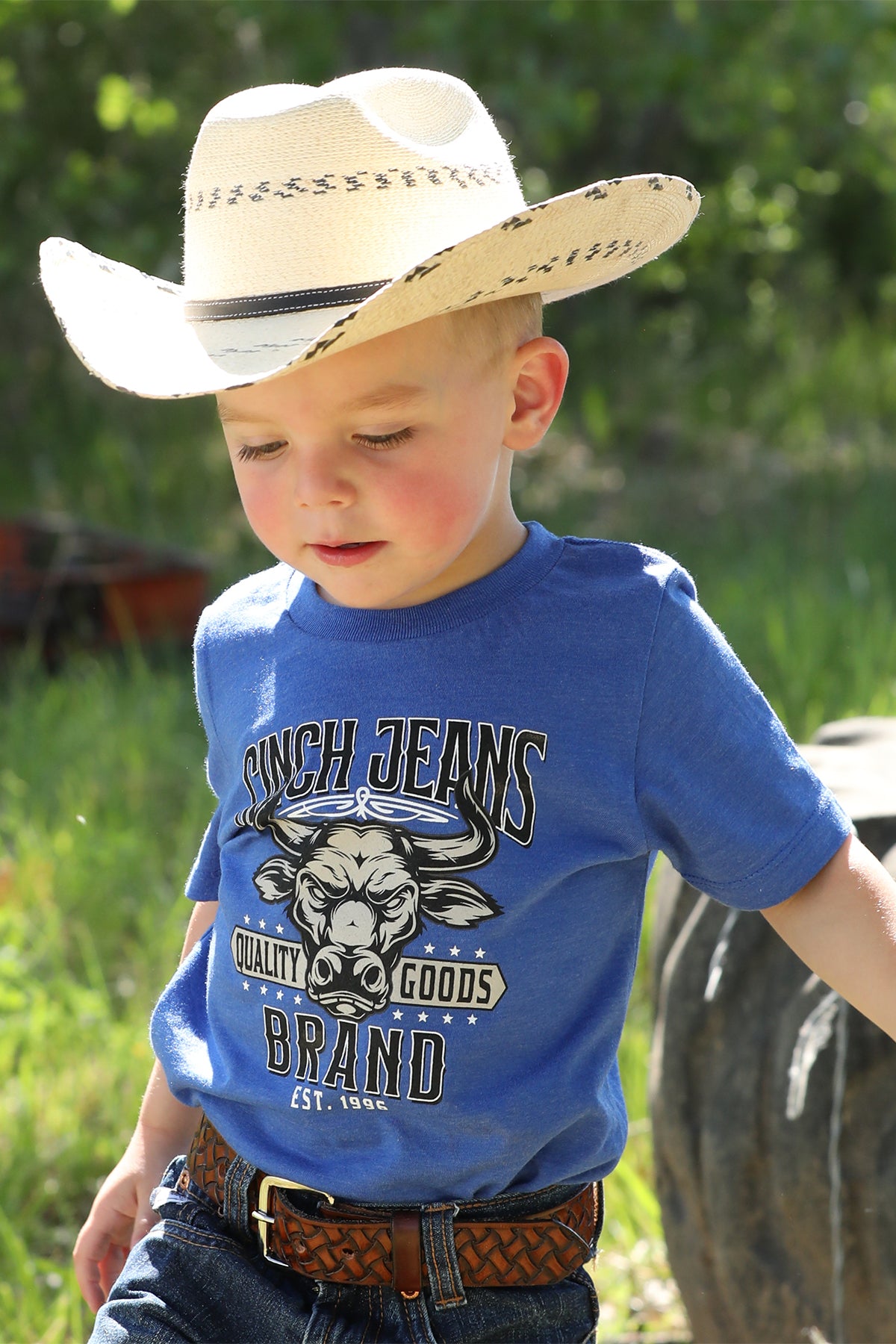 Little boy standing outside wearing a blue t-shirt with jeans, a belt, and a cowboy hat