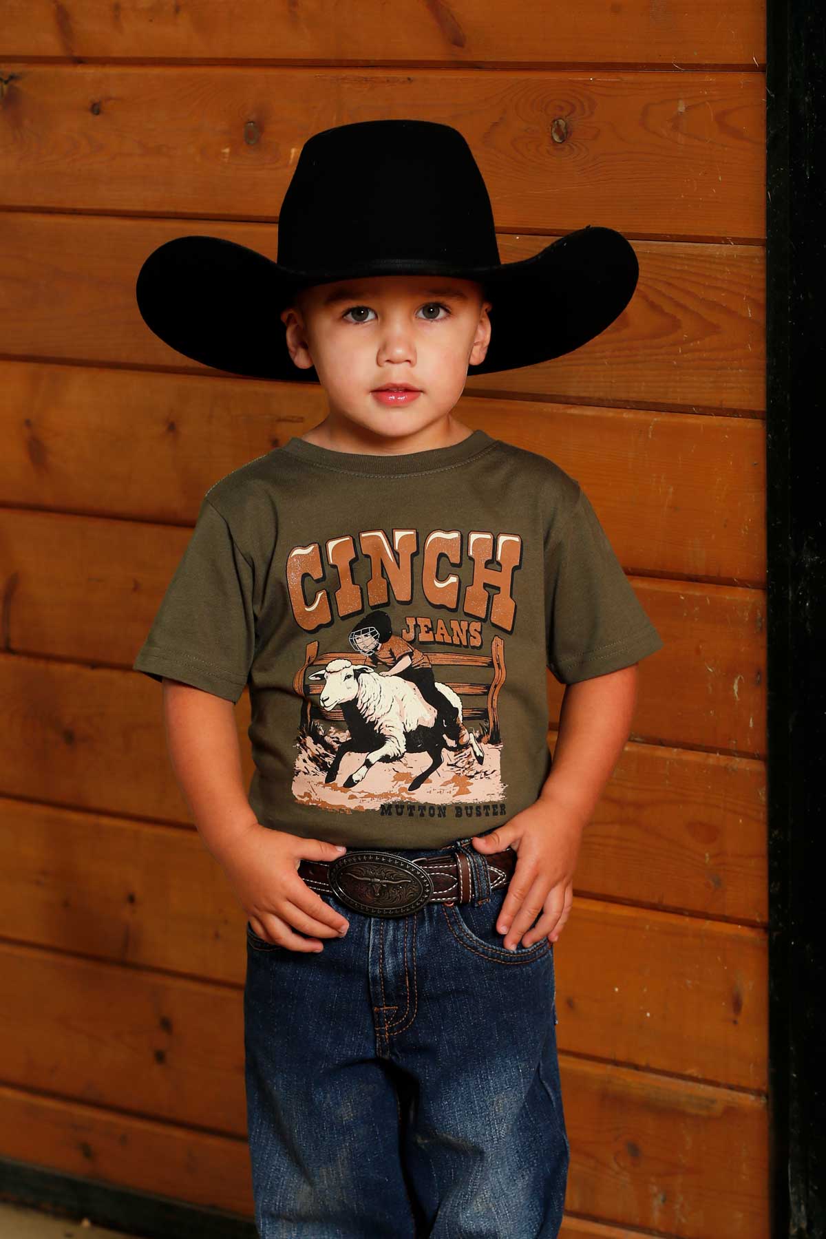 Young boy wearing a green 'CINCH' t-shirt with jeans and a cowboy hat standing in front of a wooden wall