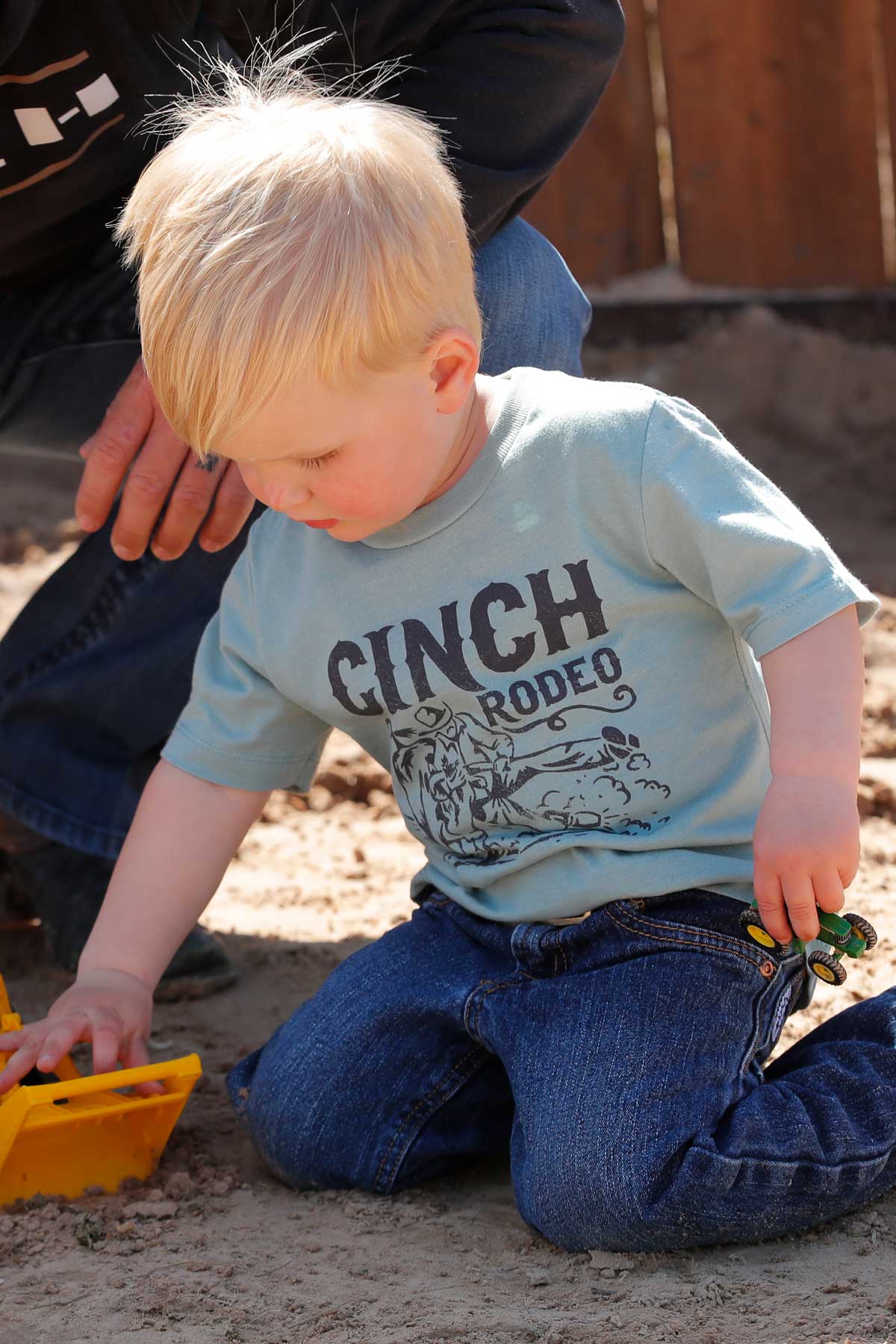 Little boy playing with toy trucks outside, wearing a green 'CINCH' t-shirt and jeans