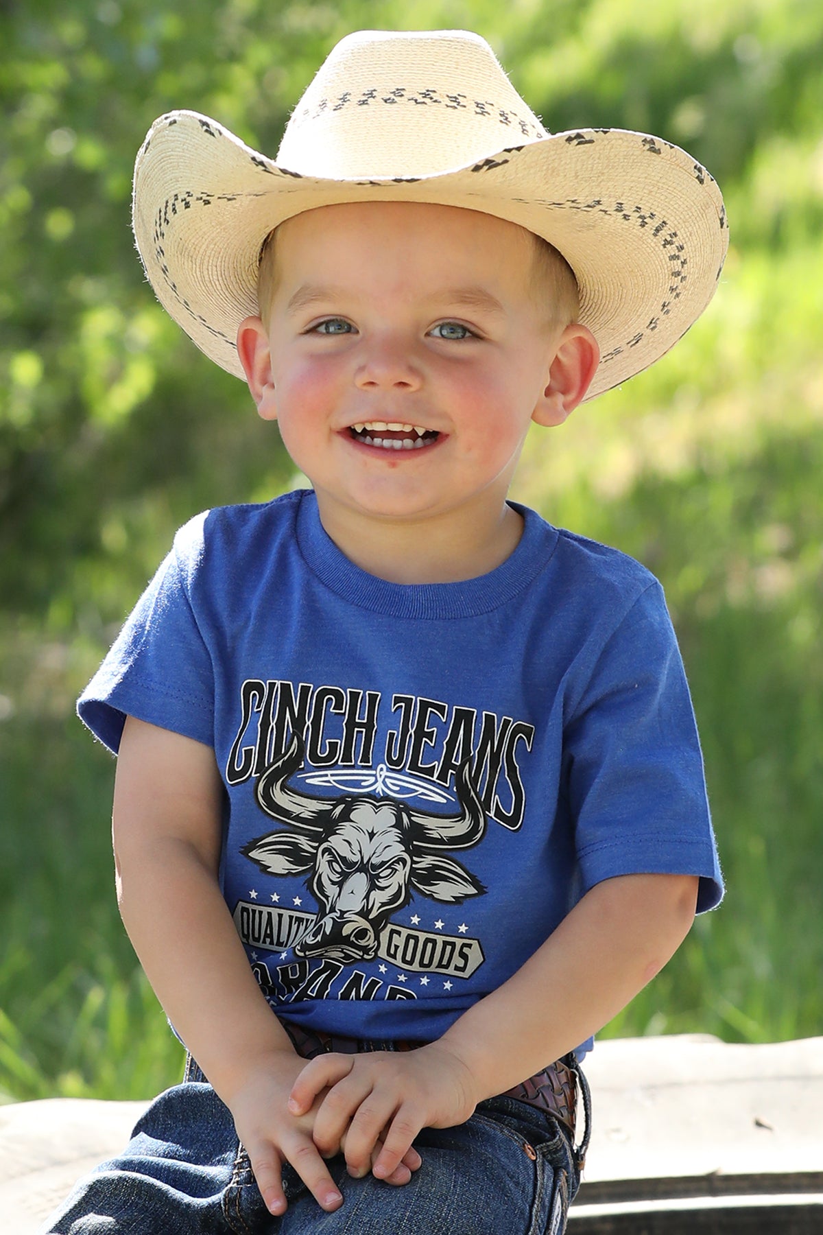 Little boy sitting outside wearing a blue t-shirt with jeans and a cowboy hat