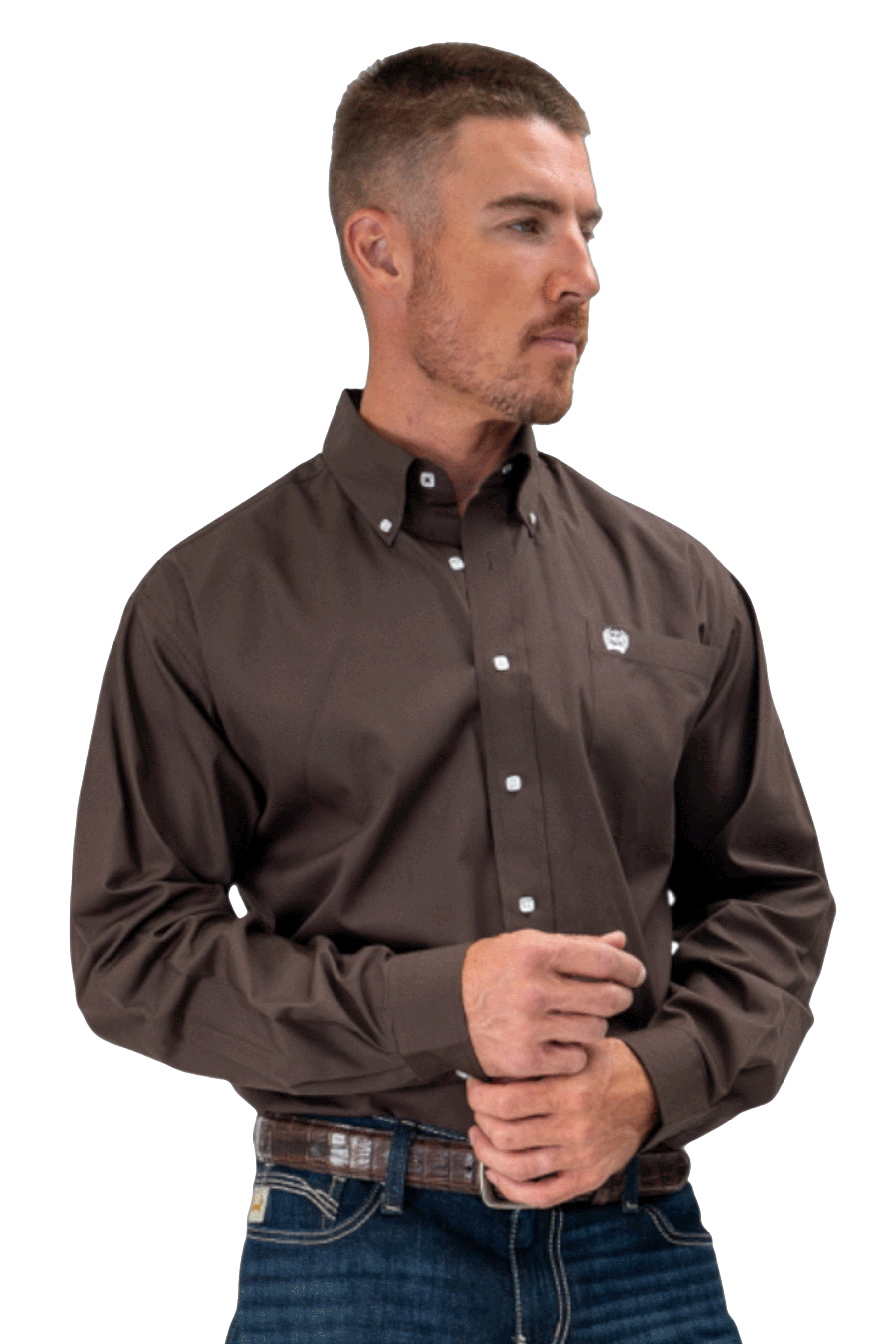 A man wearing a dark brown button-up shirt and jeans looks to the side with his hands clasped in front of him