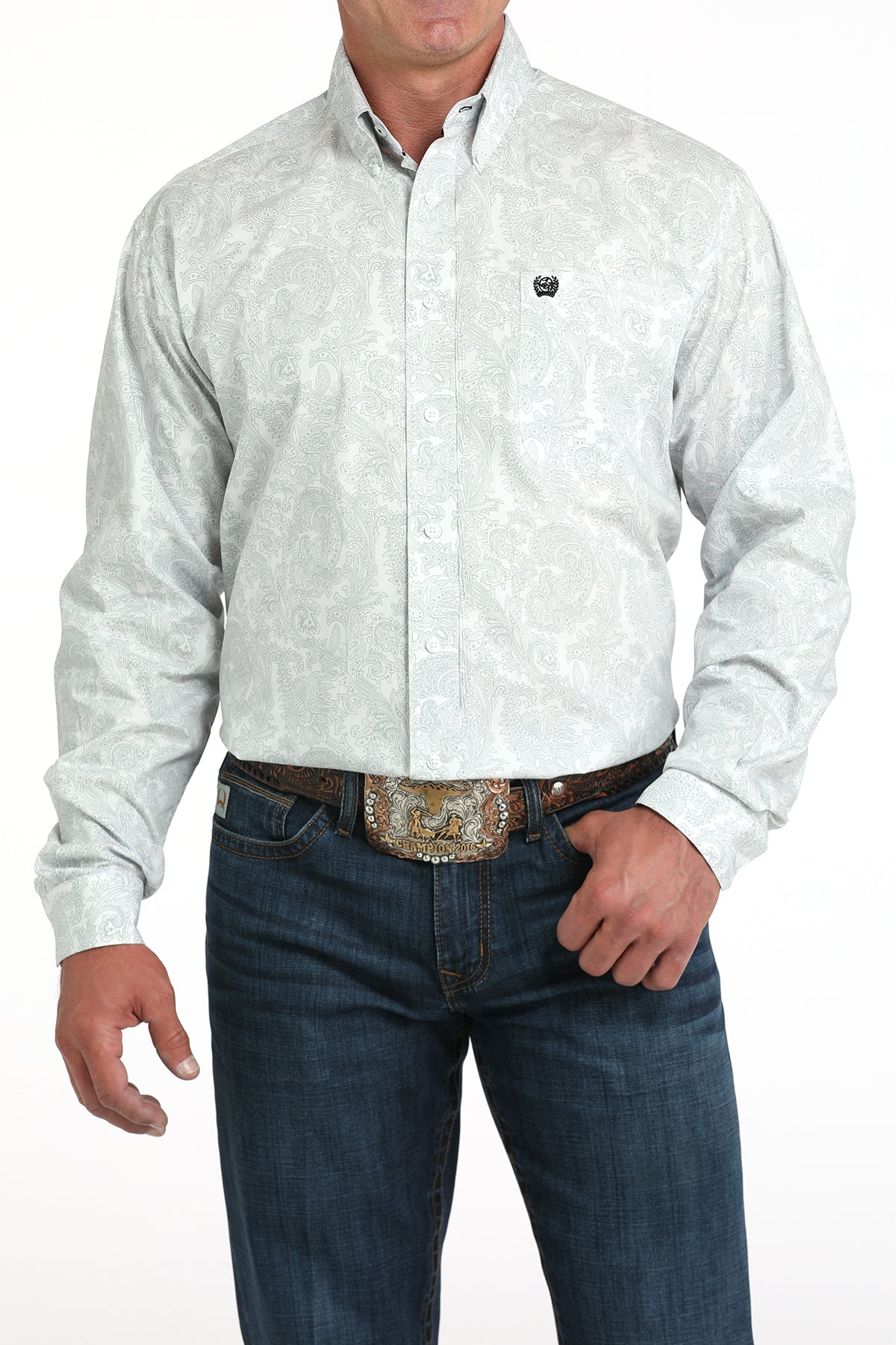 A man wearing a light patterned button-up shirt, dark jeans, and a large belt buckle stands against a white background