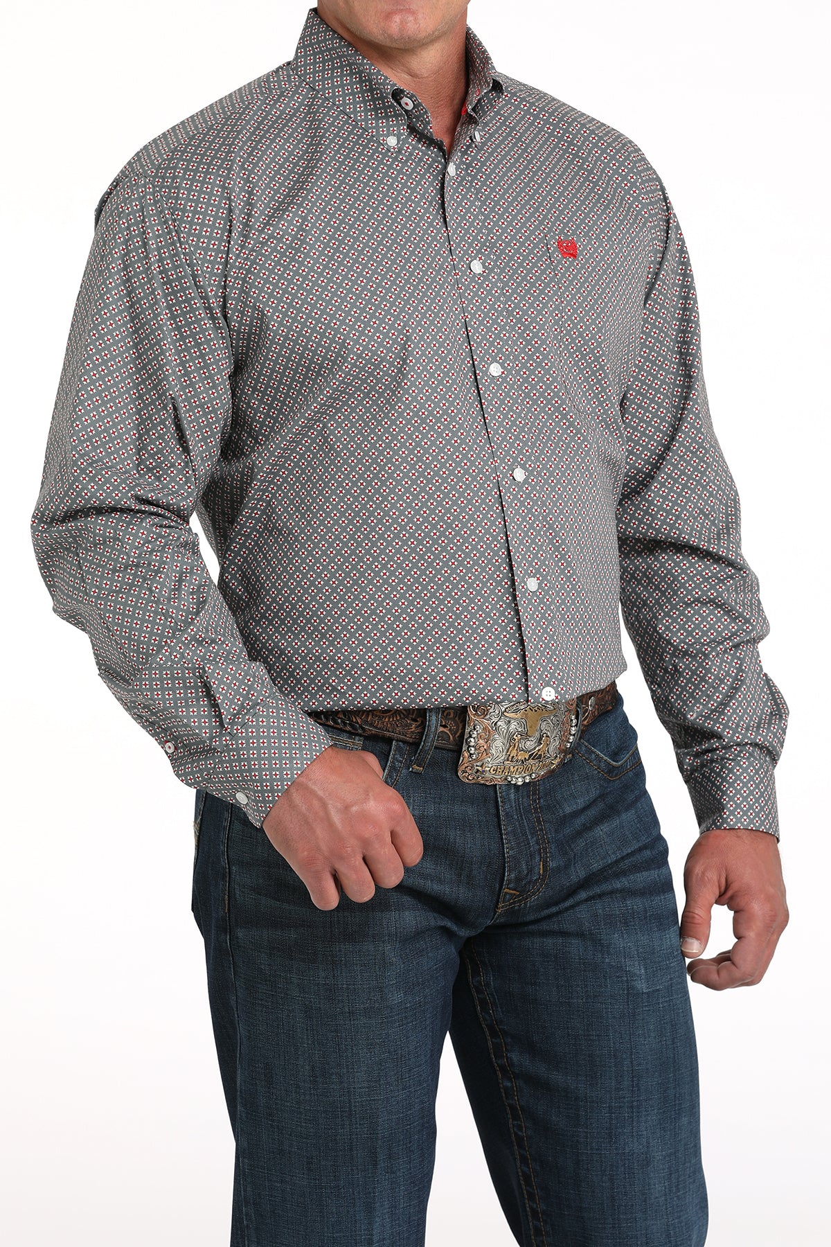 A man wearing a gray patterned button-up shirt, dark jeans, and a large belt buckle stands against a white background