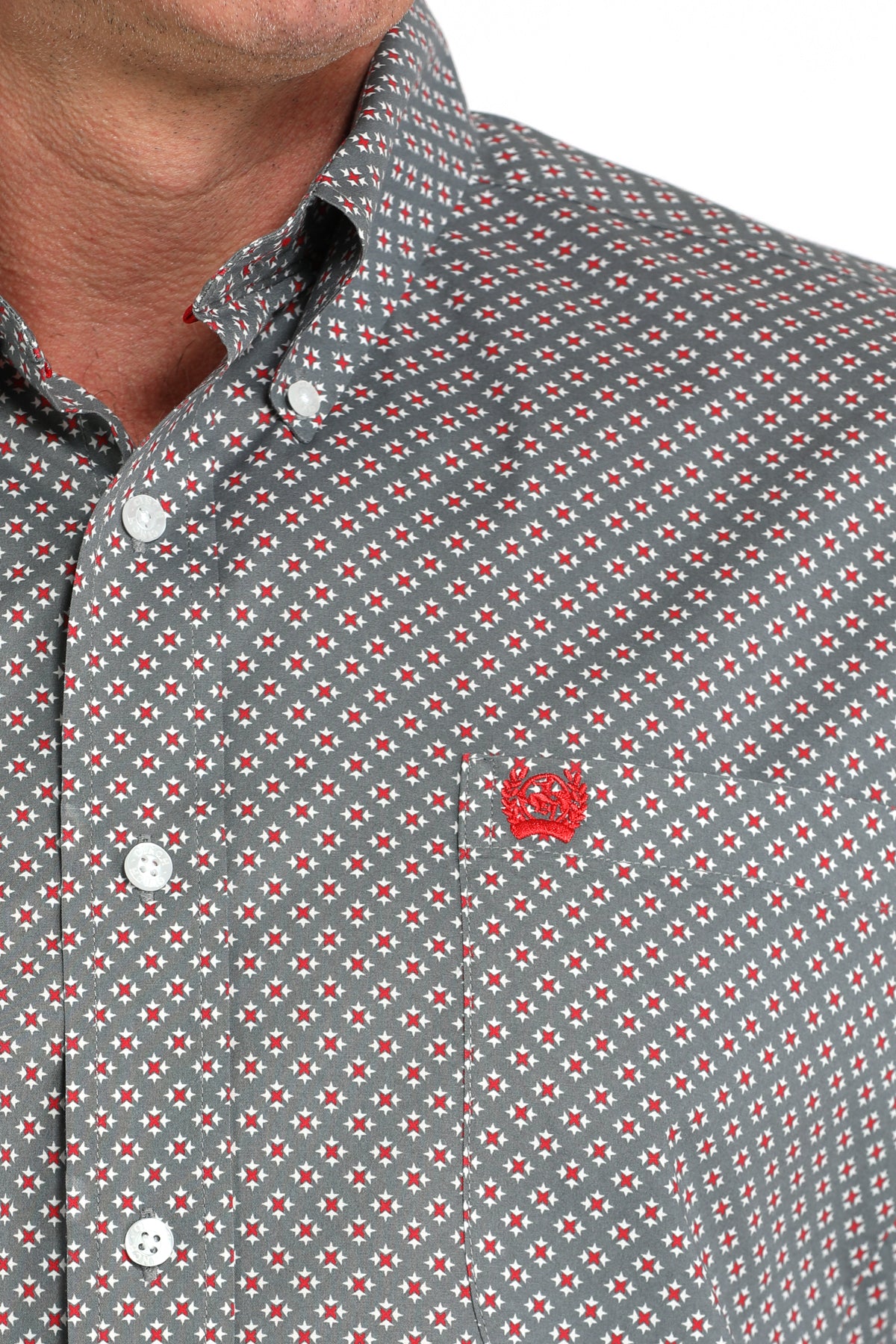 Close-up of a man wearing a gray patterned button-up shirt with a small red emblem on the chest, displayed against a white background
