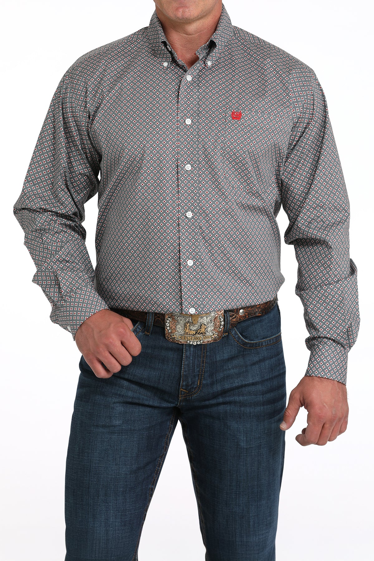 A man wearing a gray patterned button-up shirt, dark jeans, and a large belt buckle stands against a white background