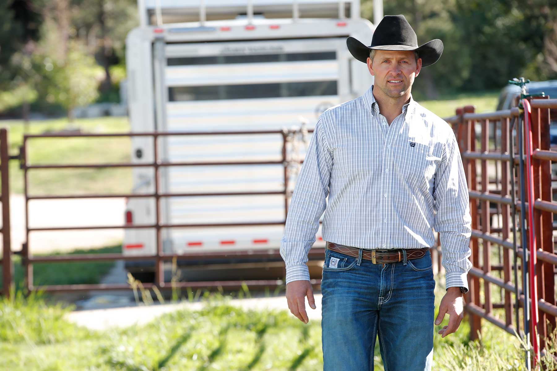 A man wearing a black cowboy hat, striped button-up shirt, jeans, and a belt stands outdoors near a metal fence and a trailer