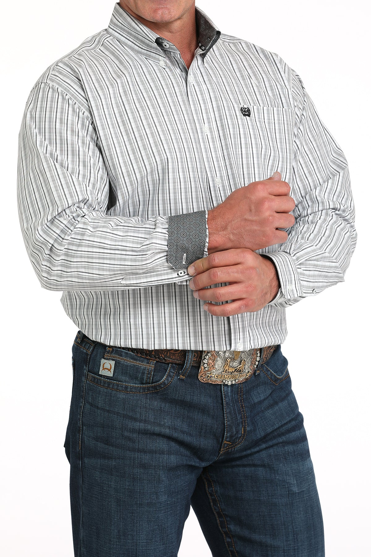 A man wearing a light patterned button-up shirt with contrasting cuffs, dark jeans, and a large belt buckle stands against a white background