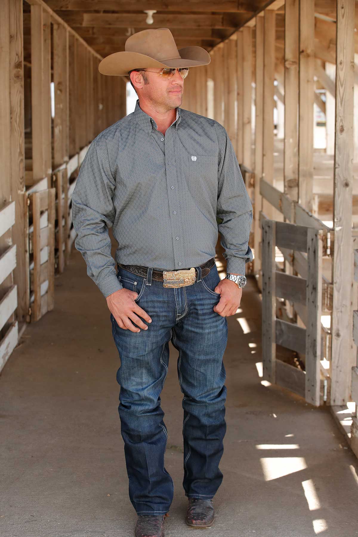 A man wearing a gray patterned button-up shirt, dark jeans, and a large belt buckle, standing in a barn