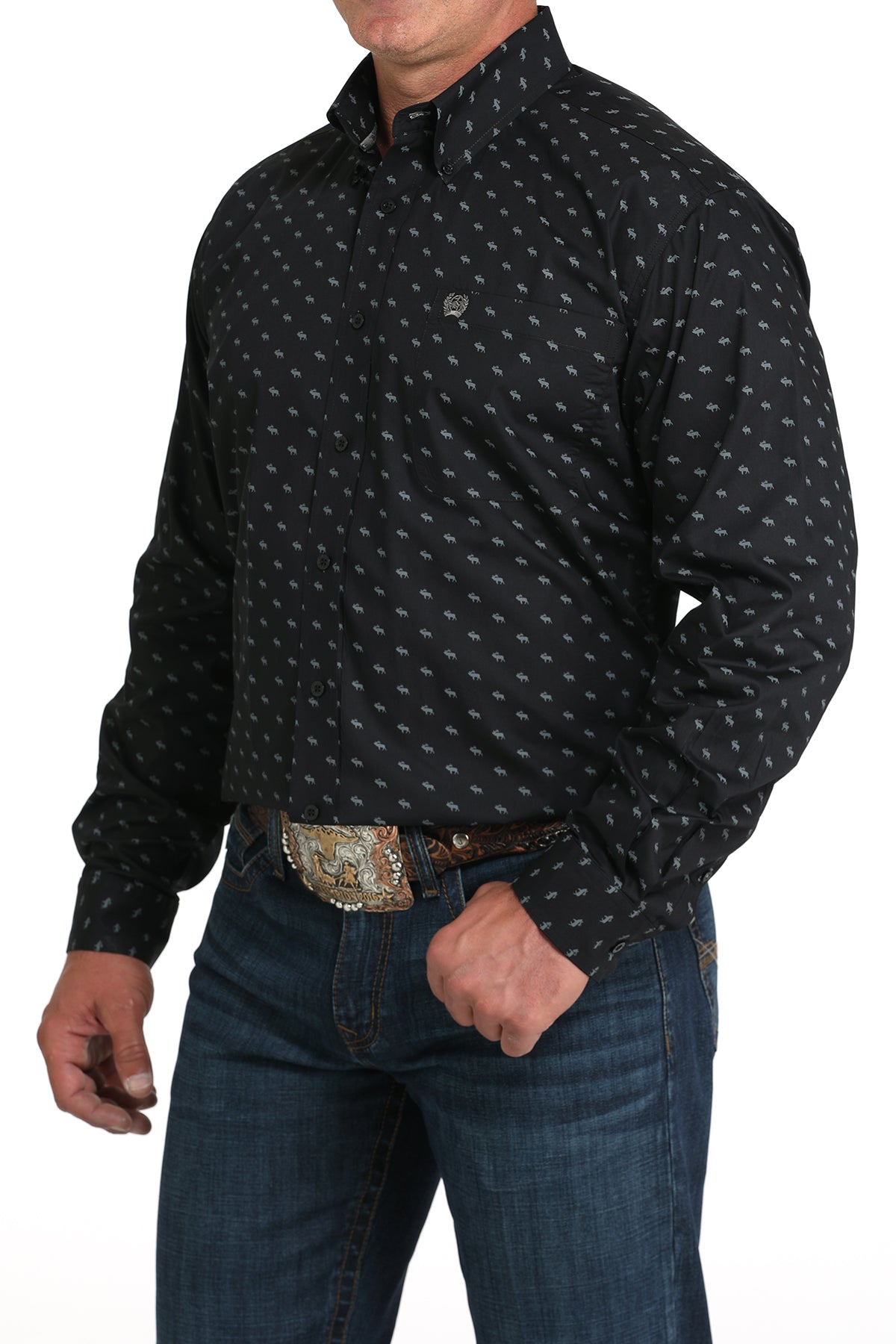 A man wearing a black patterned button-up shirt, dark jeans, and a large belt buckle, standing against a white background