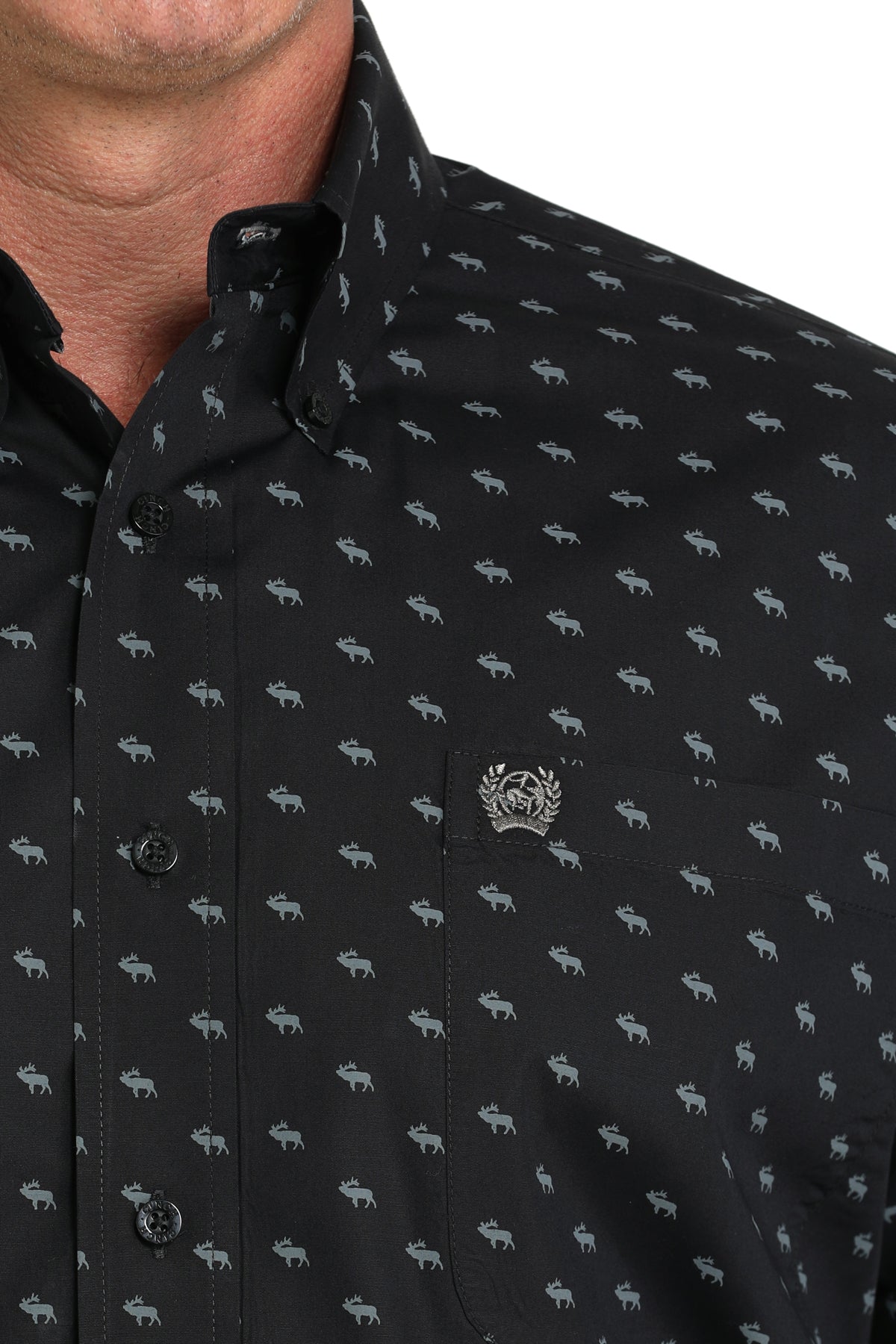Close-up of a man wearing a black patterned button-up shirt with a small emblem on the chest, displayed against a white background