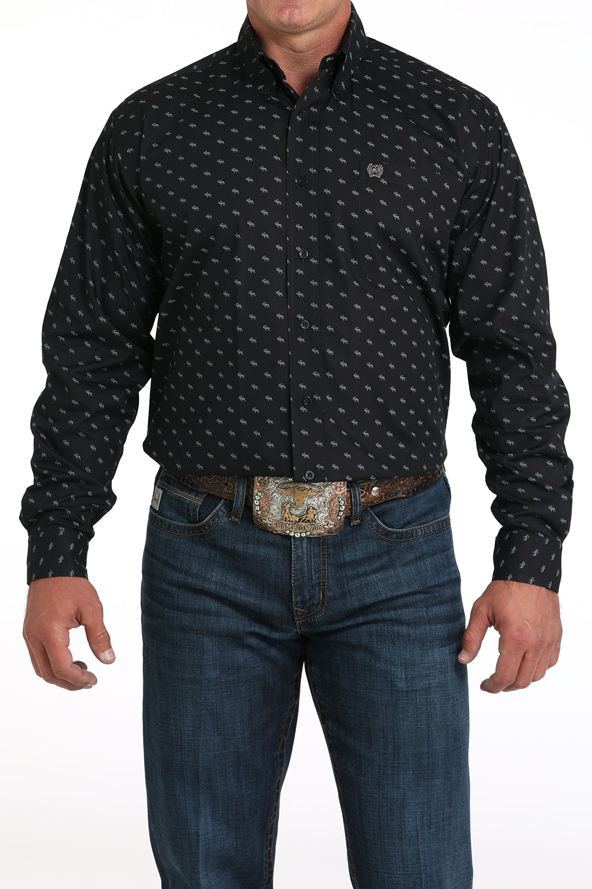 A man wearing a black patterned button-up shirt, dark jeans, and a large belt buckle, standing against a white background