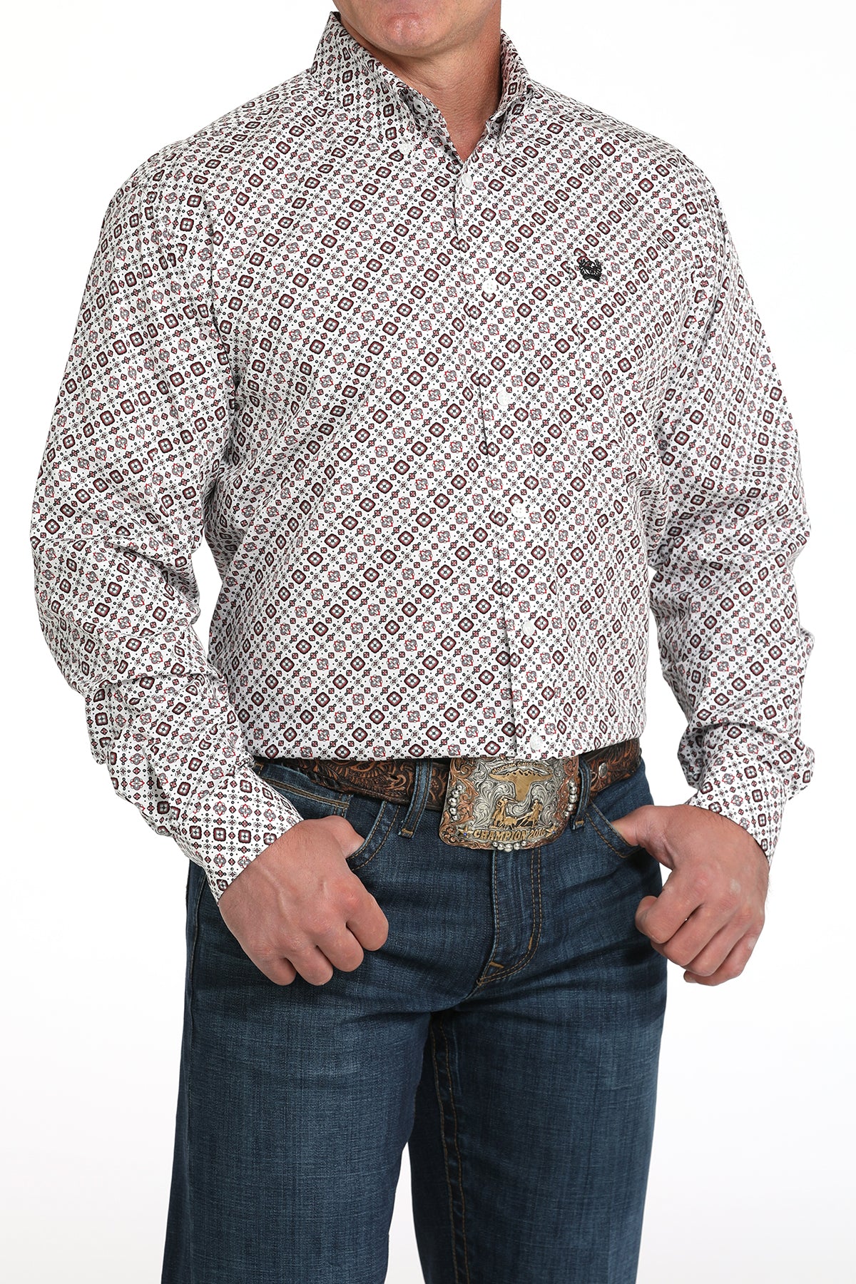 A man wearing a light patterned button-up shirt, dark jeans, and a large belt buckle, standing against a white background