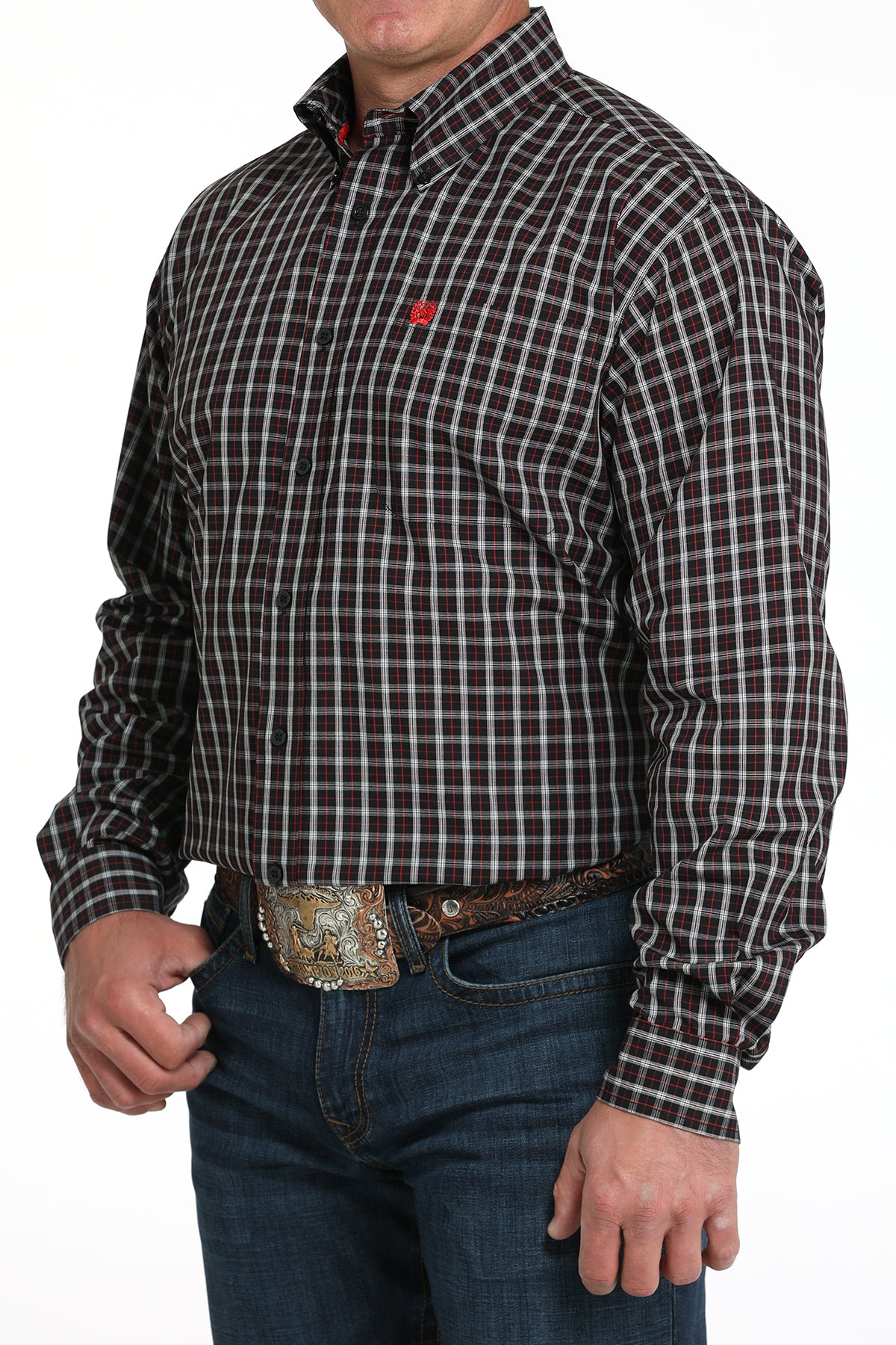 A man wearing a black patterned button-up shirt, dark jeans, and a large belt buckle, standing against a white background