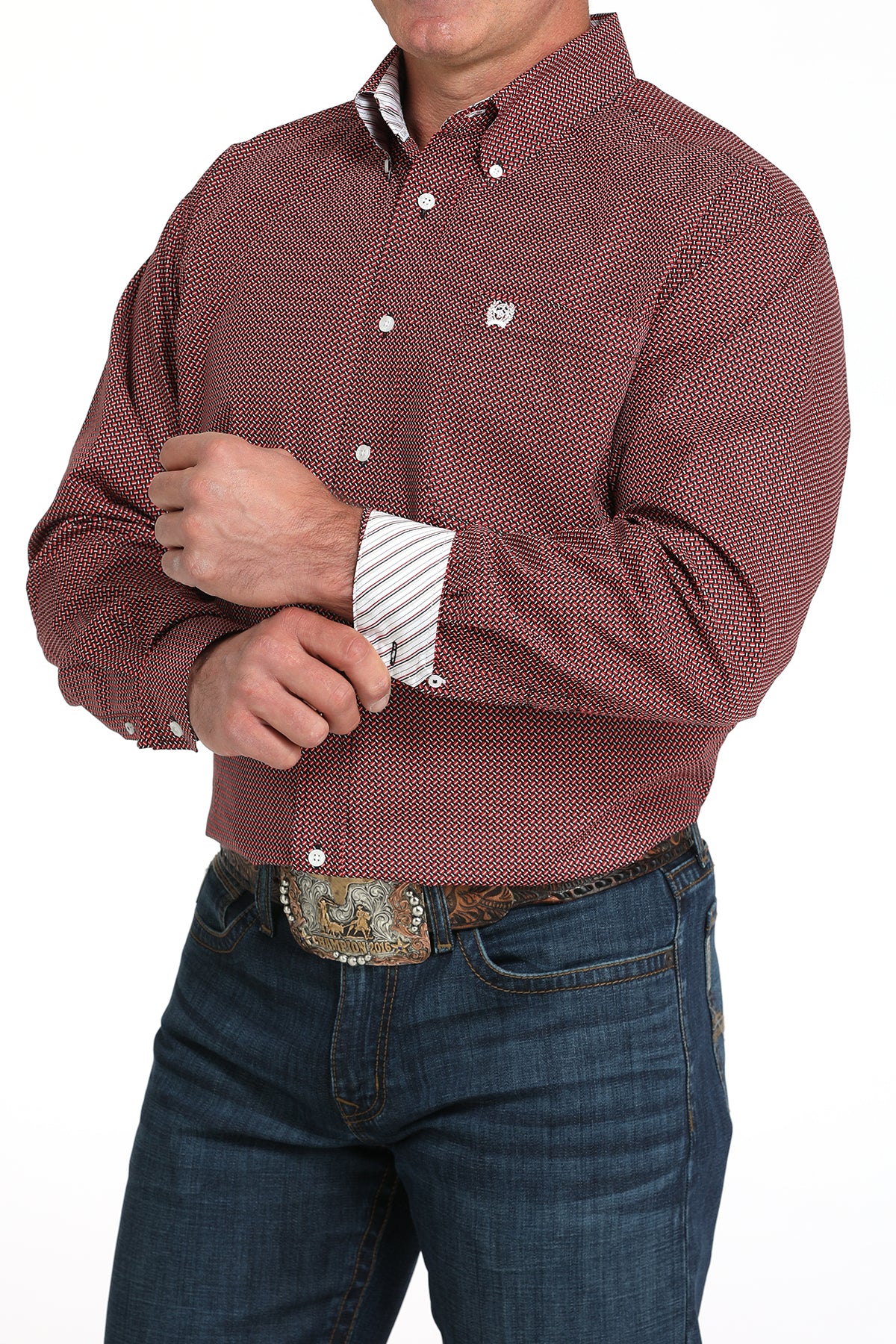 A man wearing a maroon patterned button-up shirt with contrasting cuffs, dark jeans, and a large belt buckle, standing against a white background