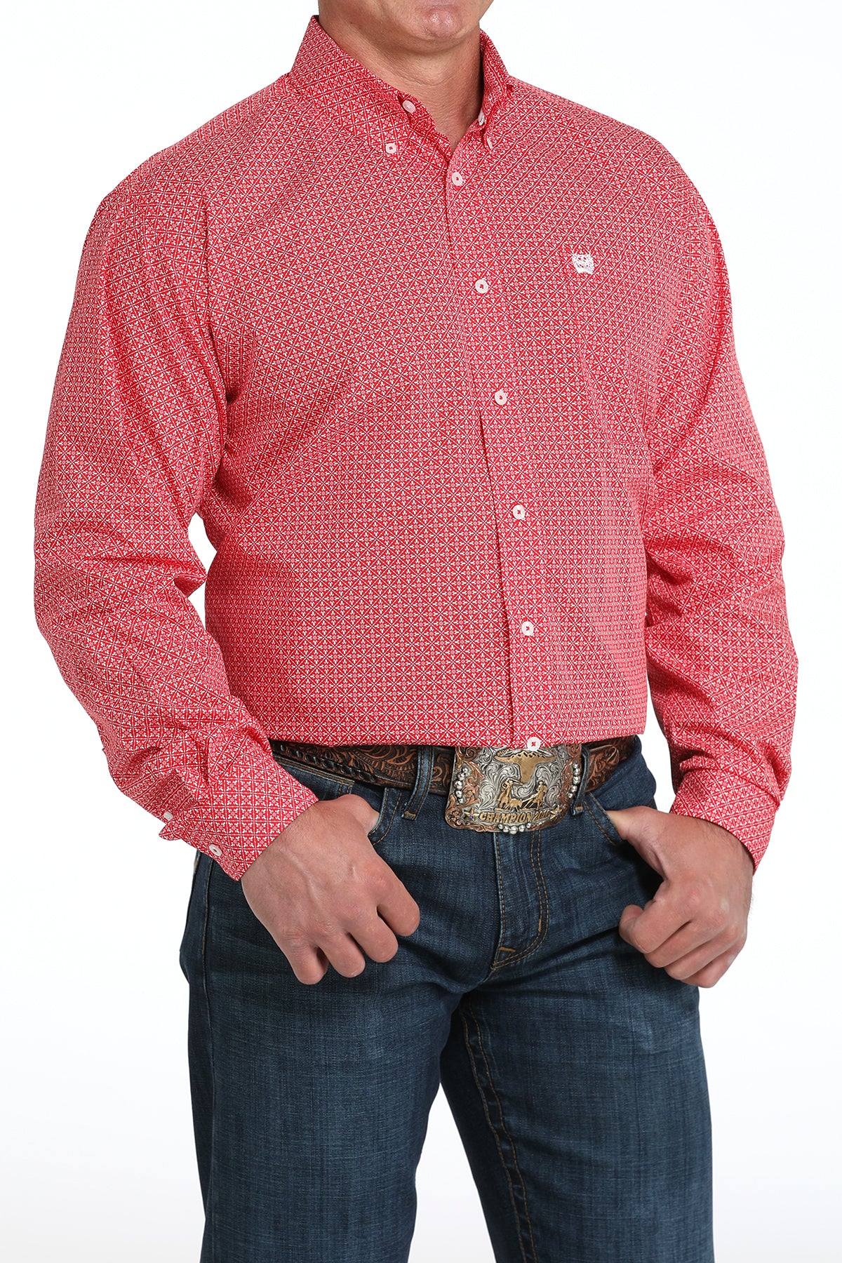 A man wearing a red patterned button-up shirt, dark jeans, and a large belt buckle, standing against a white background