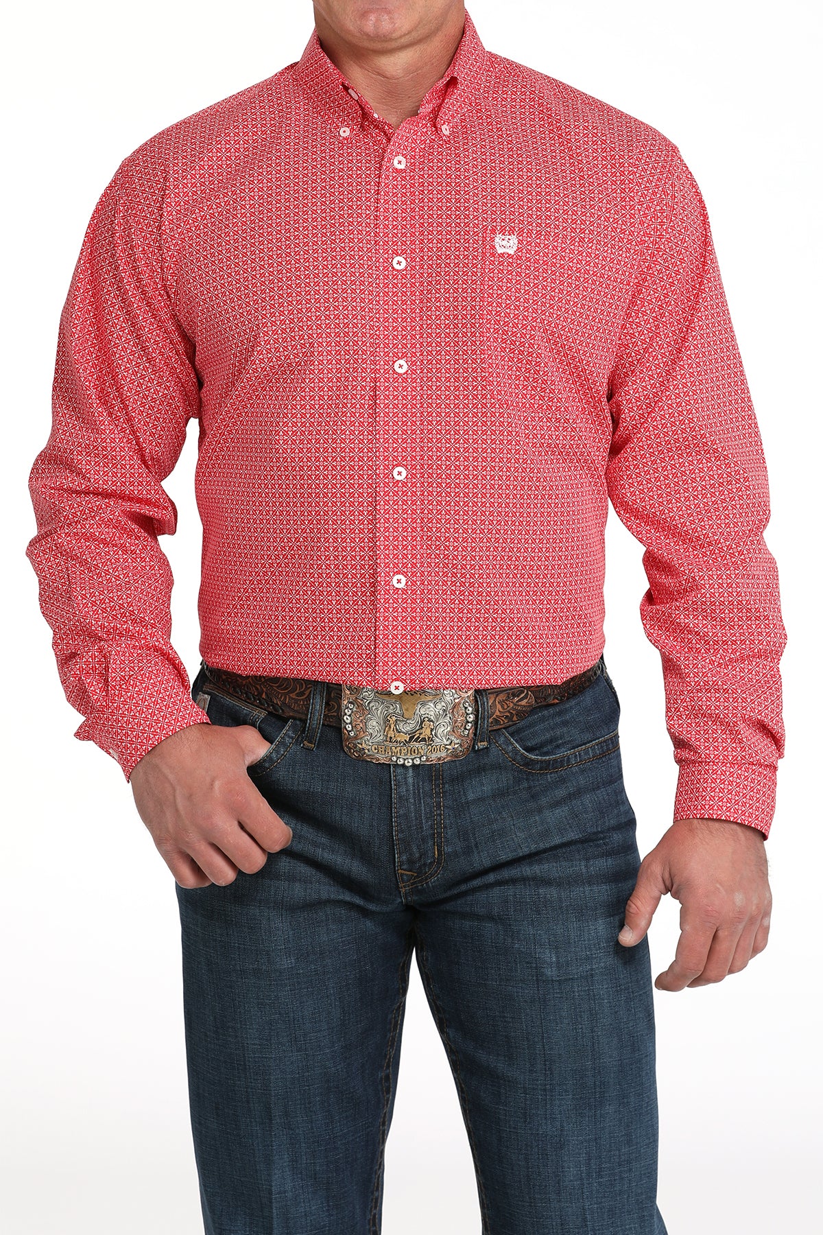 A man wearing a red patterned button-up shirt, dark jeans, and a large belt buckle, standing against a white background
