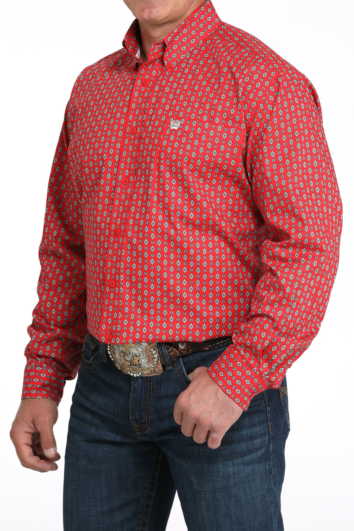 A man wearing a red patterned button-up shirt, dark jeans, and a large belt buckle, standing against a white background