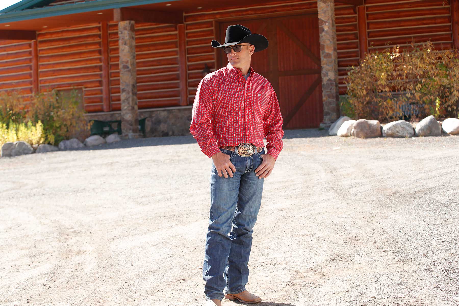 Man standing outside in front of a building, wearing a red long sleeve button-down shirt, a black cowboy hat, jeans and a belt