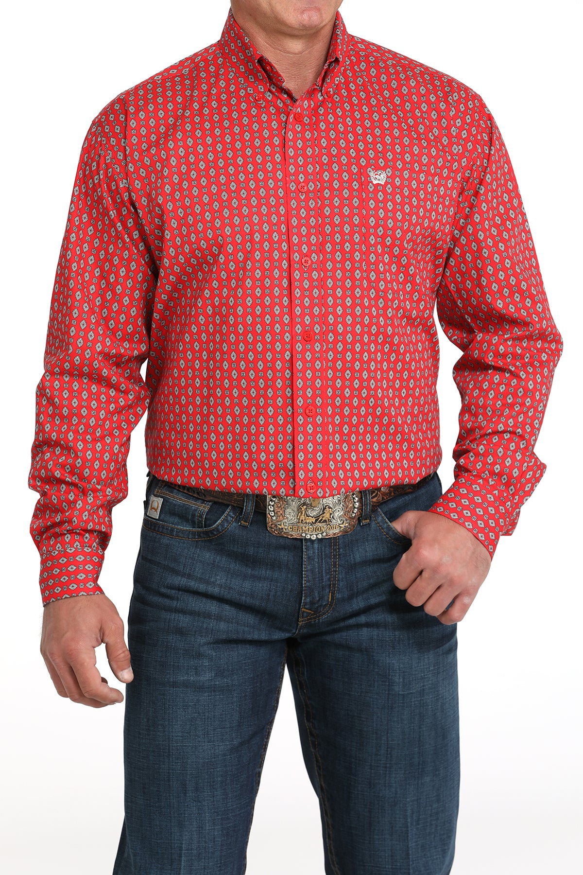 A man wearing a red patterned button-up shirt, dark jeans, and a large belt buckle, standing against a white background