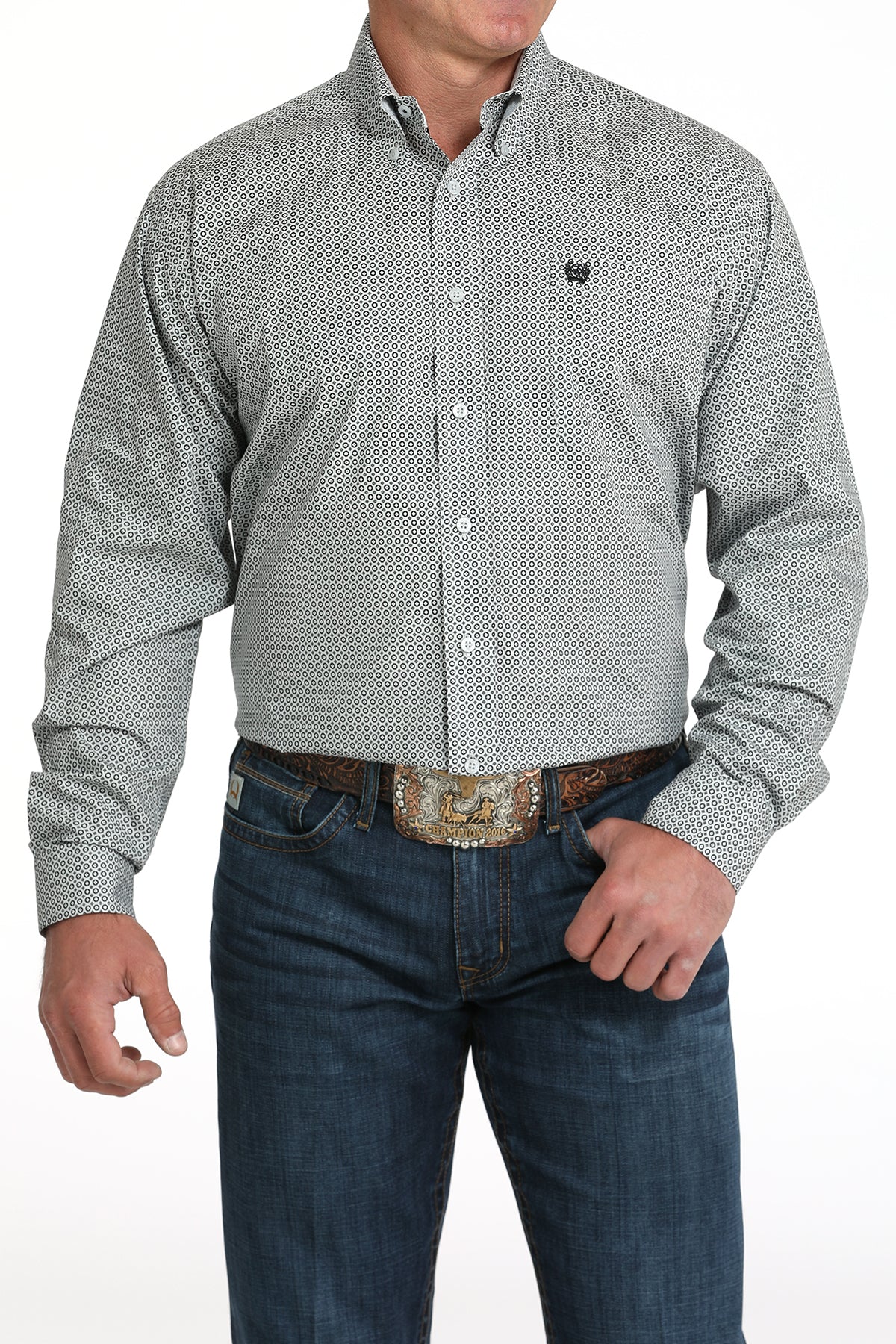 A man wearing a gray patterned button-up shirt, dark jeans, and a large belt buckle, standing against a white background