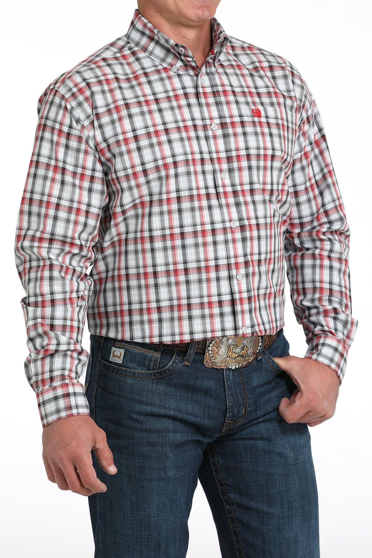 A man wearing a red, white and black plaid button-up shirt, dark jeans, and a large belt buckle, standing against a white background