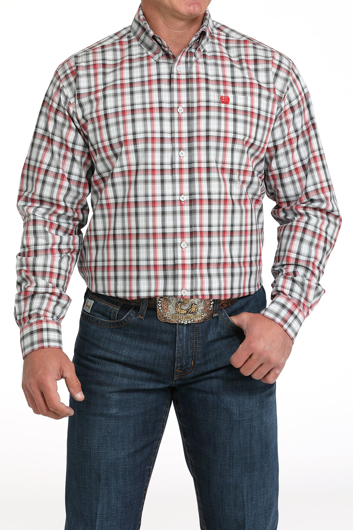 A man wearing a red, white and black plaid button-up shirt, dark jeans, and a large belt buckle, standing against a white background