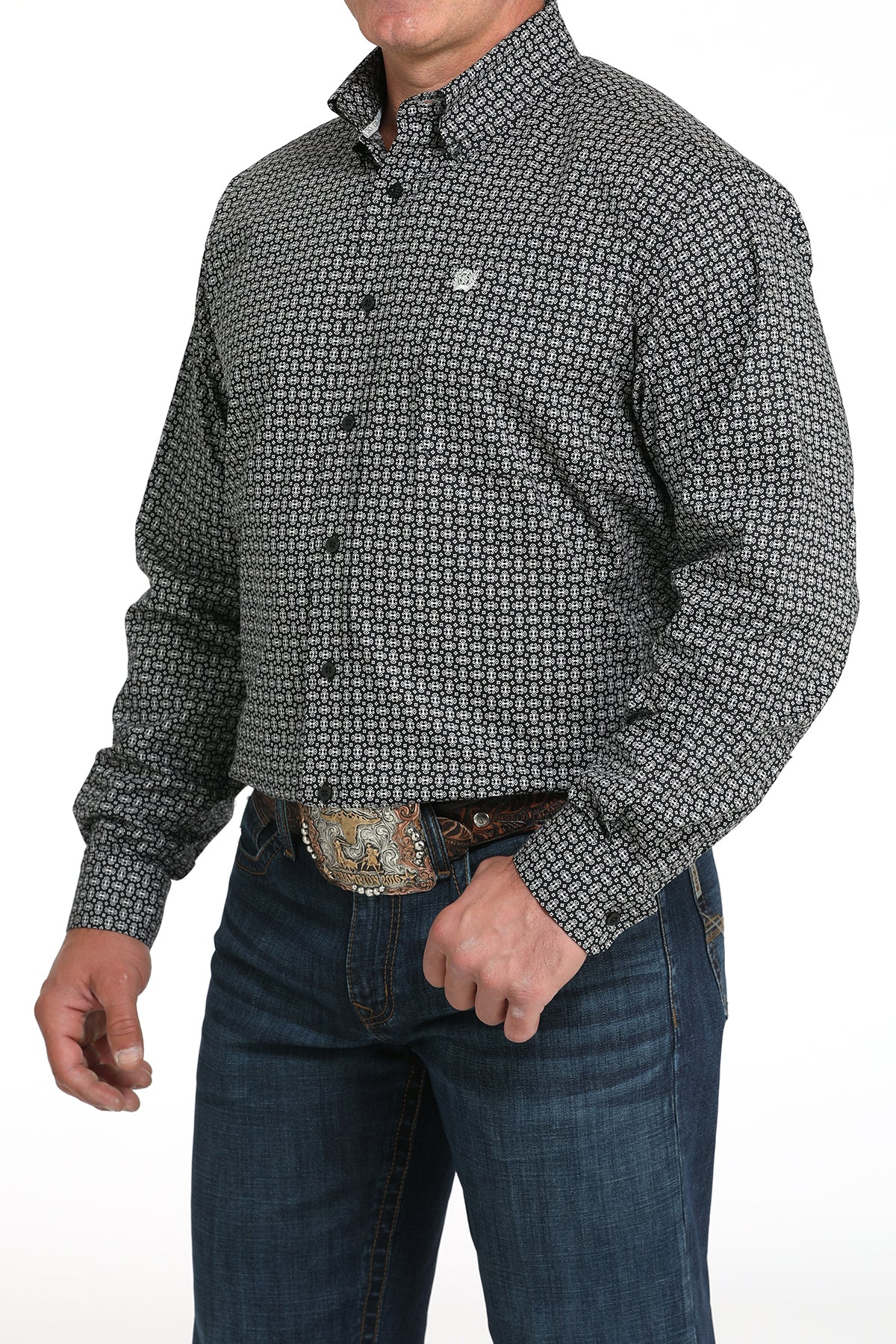 A man wearing a gray patterned button-up shirt, dark jeans, and a large belt buckle, standing against a white background