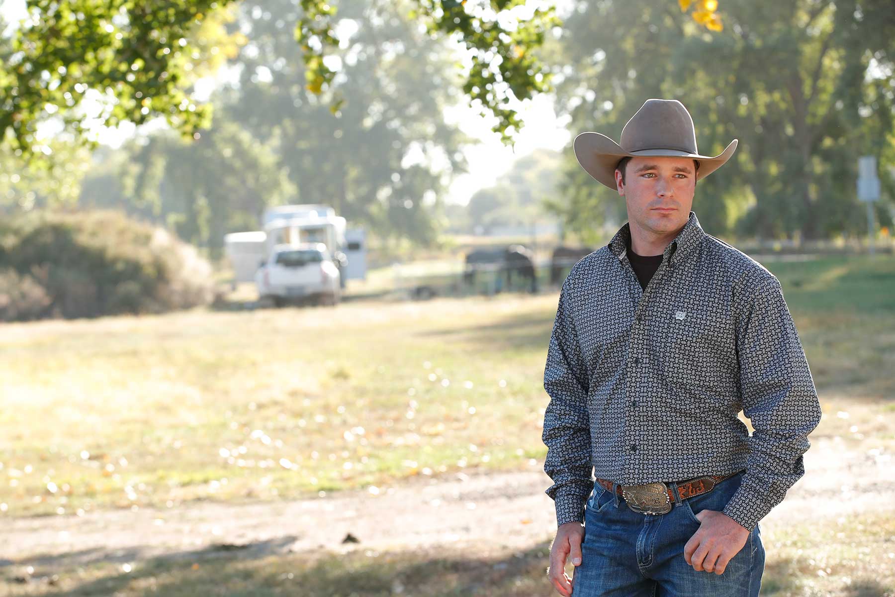 A man wearing a cowboy hat, patterned shirt, jeans, and a belt buckle stands outdoors on a sunny day with trees and trucks in the background