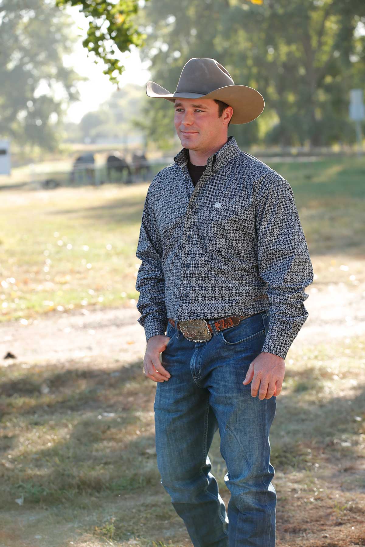 A man wearing a cowboy hat, patterned shirt, jeans, and a belt buckle stands outdoors on a sunny day with trees in the background