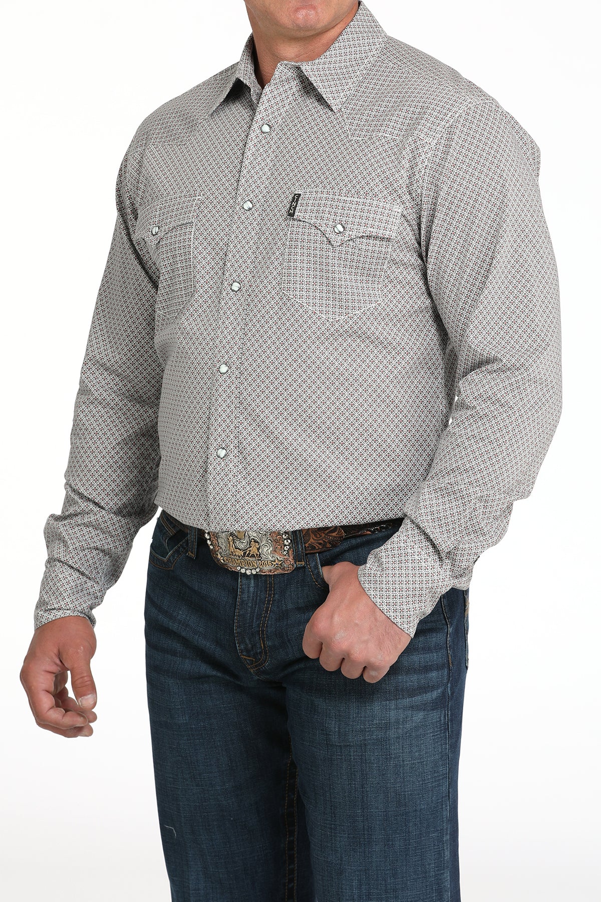 A man wearing a light gray patterned button-up shirt, dark jeans, and a large belt buckle, standing against a white background