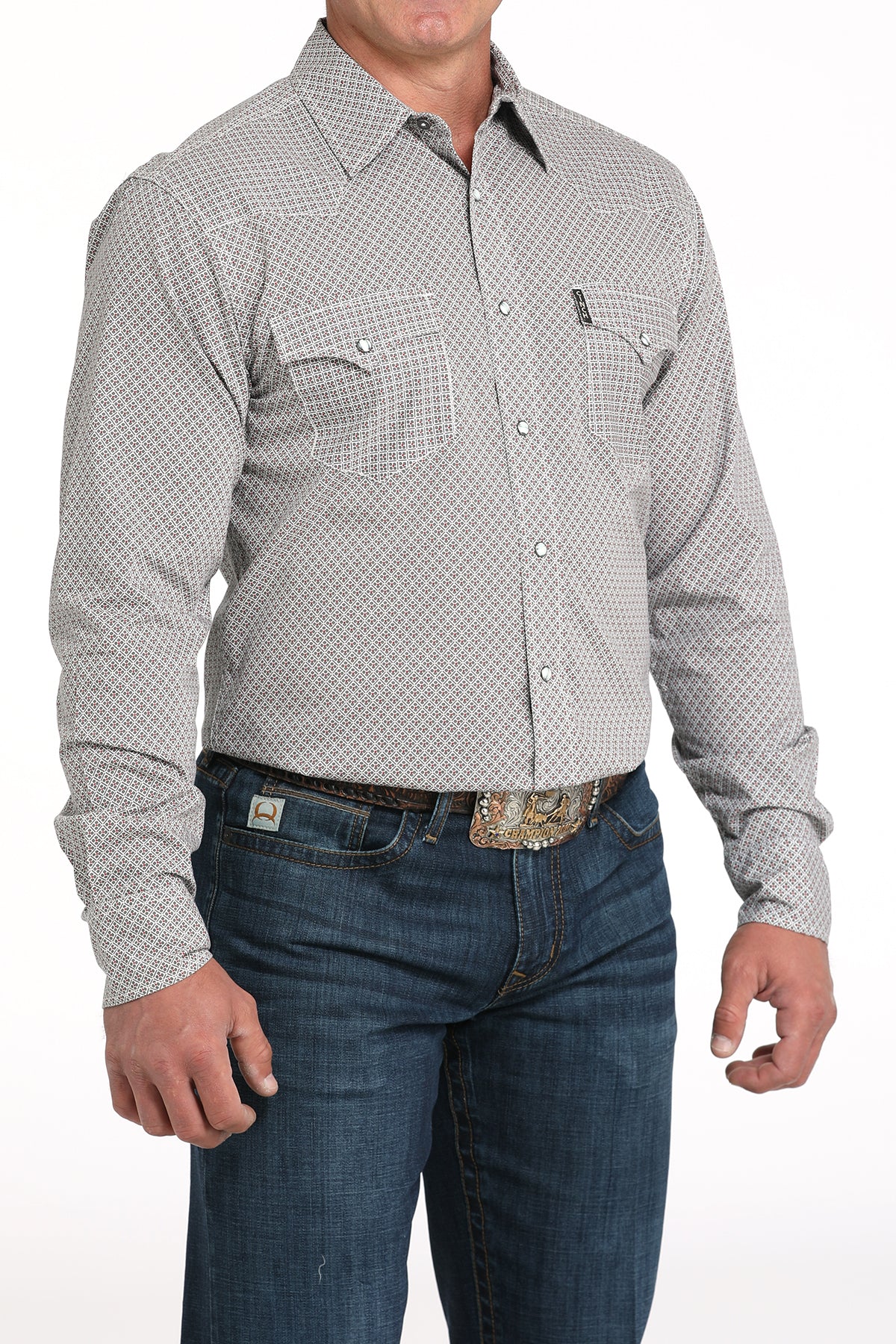 A man wearing a light gray patterned button-up shirt, dark jeans, and a large belt buckle, standing against a white background