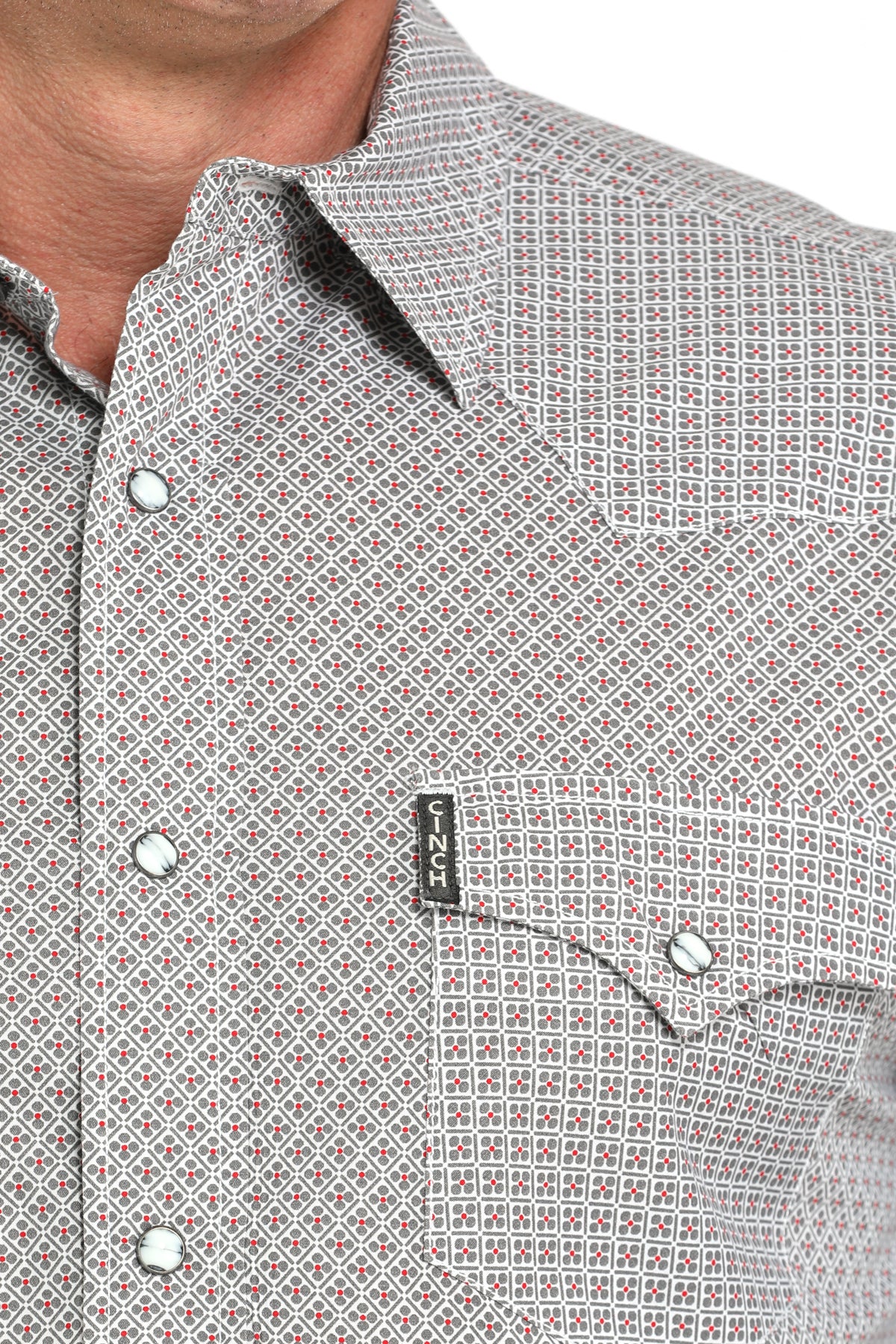 Close-up of a man wearing a light gray patterned button-up shirt with a small emblem on the chest, displayed against a white background