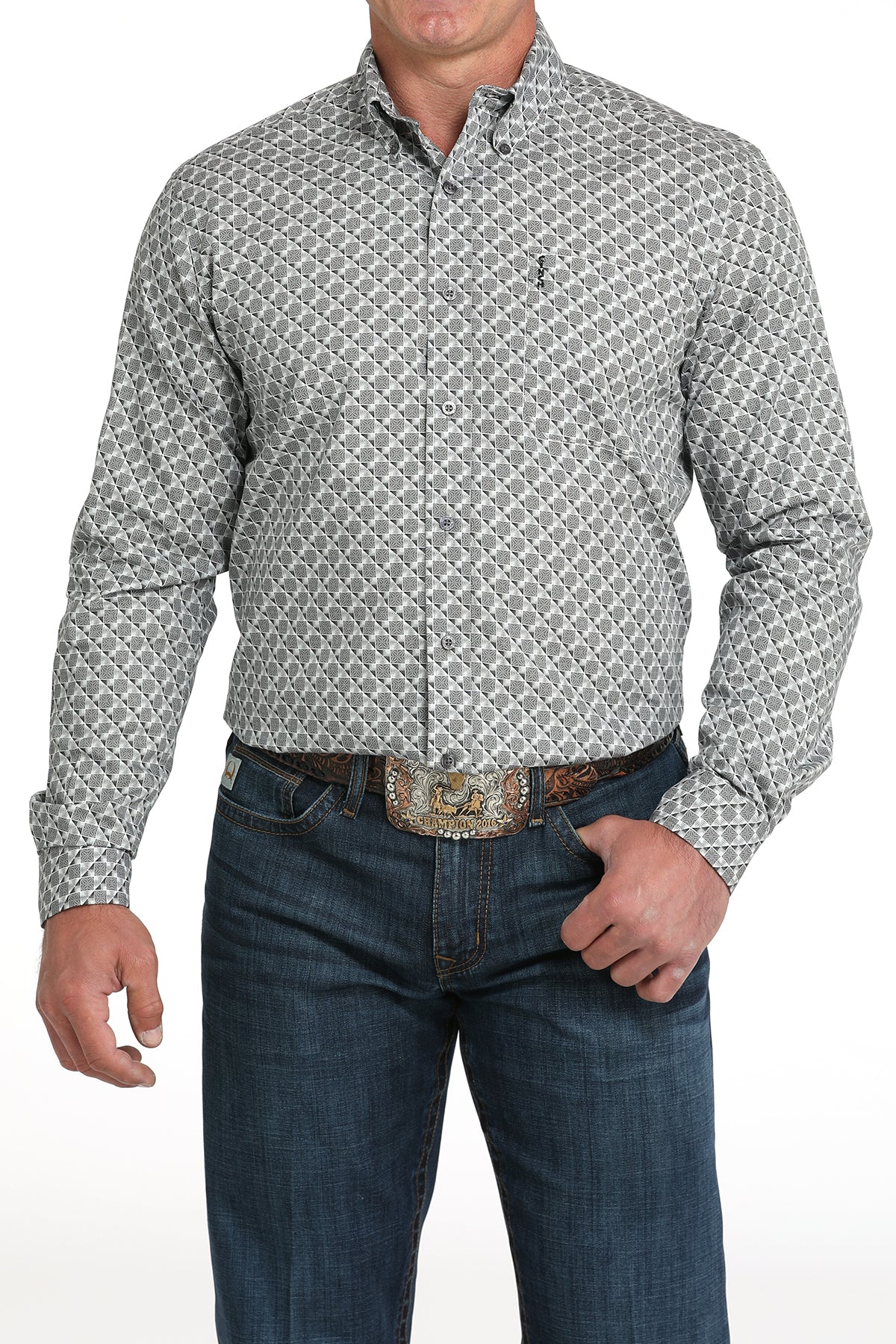 A man wearing a light gray patterned button-up shirt, dark jeans, and a large belt buckle, standing against a white background