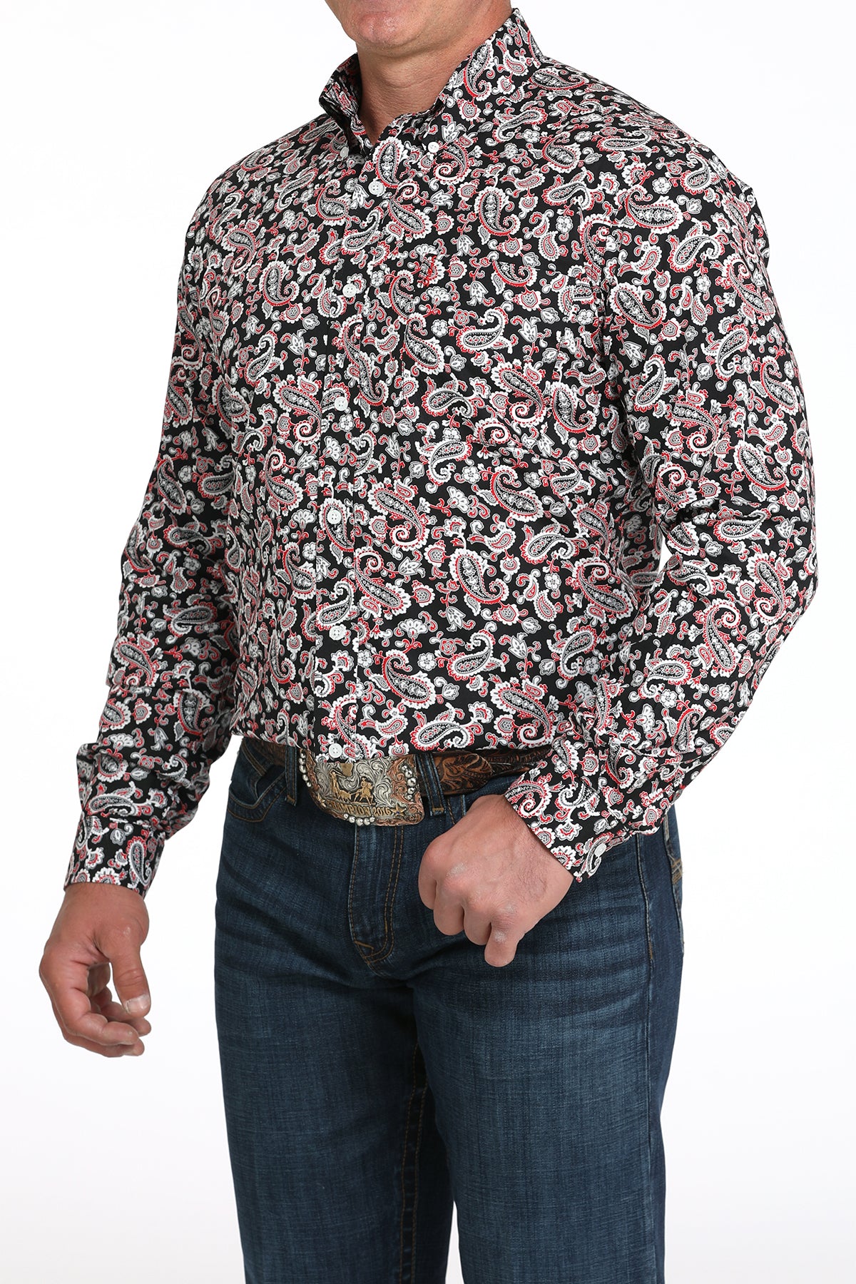 A man wearing a red and black patterned button-up shirt, dark jeans, and a large belt buckle, standing against a white background