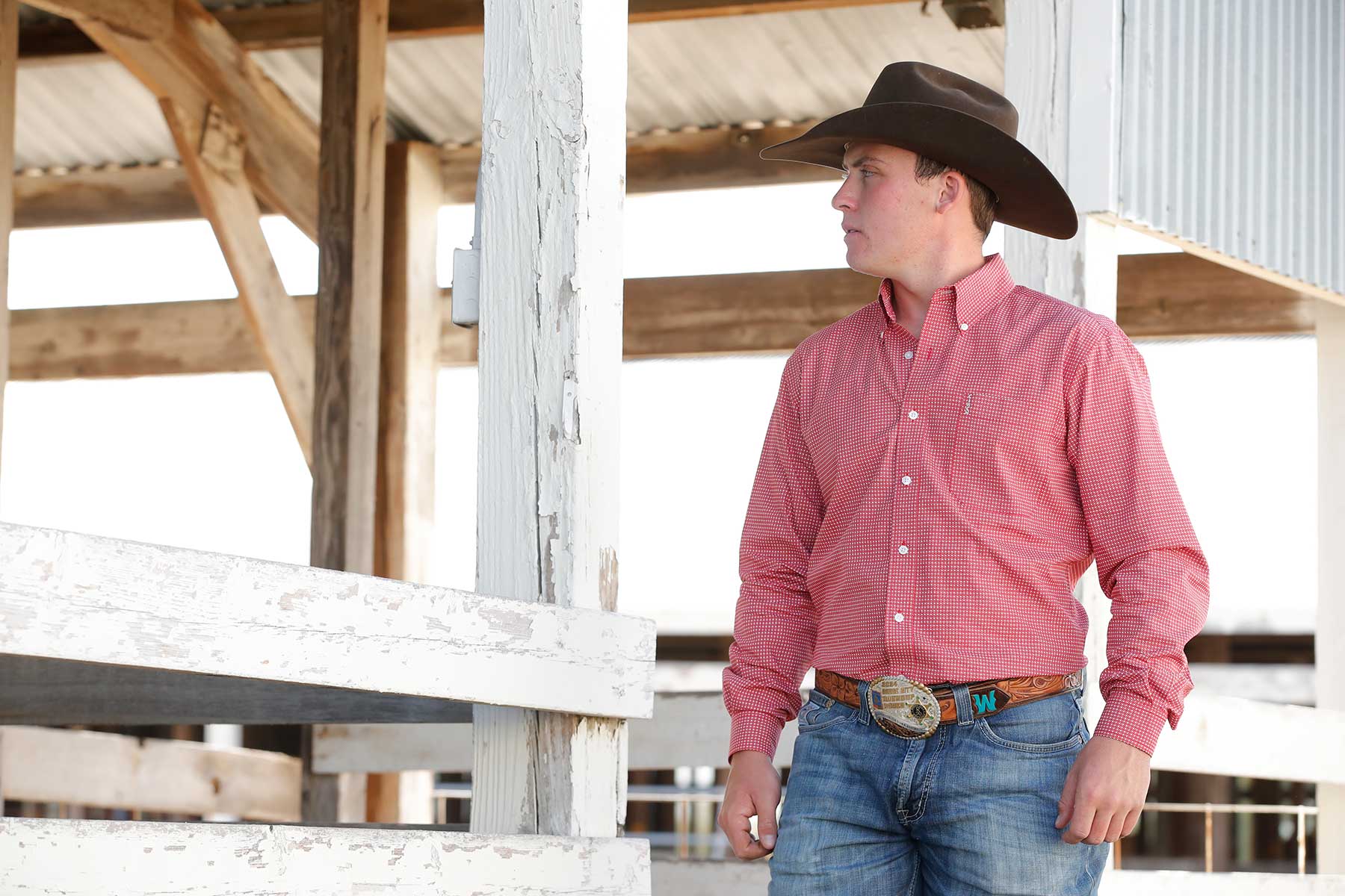Man wearing a black cowboy hat, red button-up shirt with a small check pattern, blue jeans, and a large belt buckle. He is standing near a white wooden fence in a rustic barn setting, looking off to the side