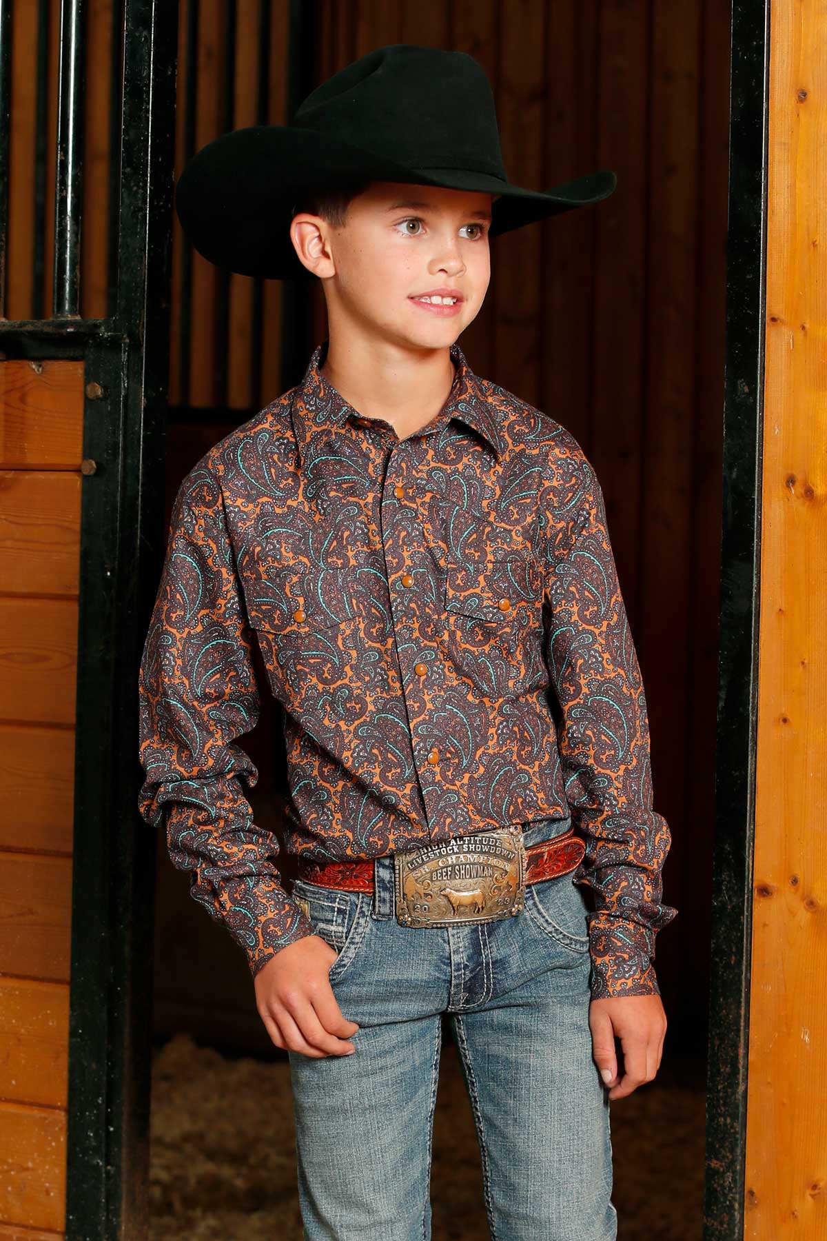 Young boy standing in a barn wearing a floral long sleeve button-down with jeans and a cowboy hat