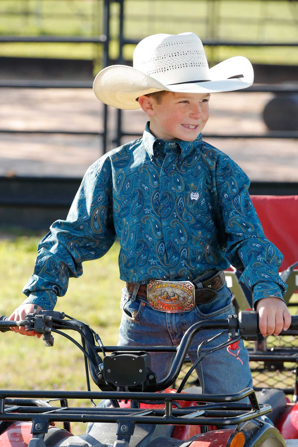 Boy who is outside sitting on a four-wheeler, wearing a blue long sleeve button down shirt, jeans, a belt, and a cowboy hat