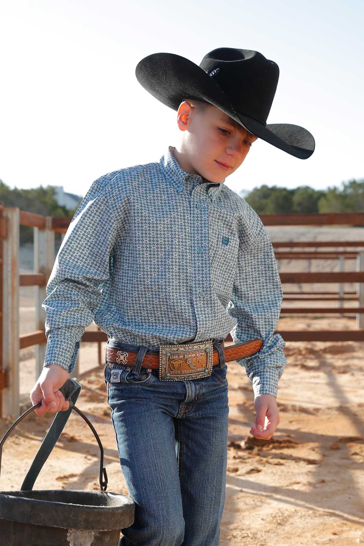Boy standing outside in an arena wearing a green long sleeve button down shirt, jeans, a belt, and a cowboy hat, holding a bucket