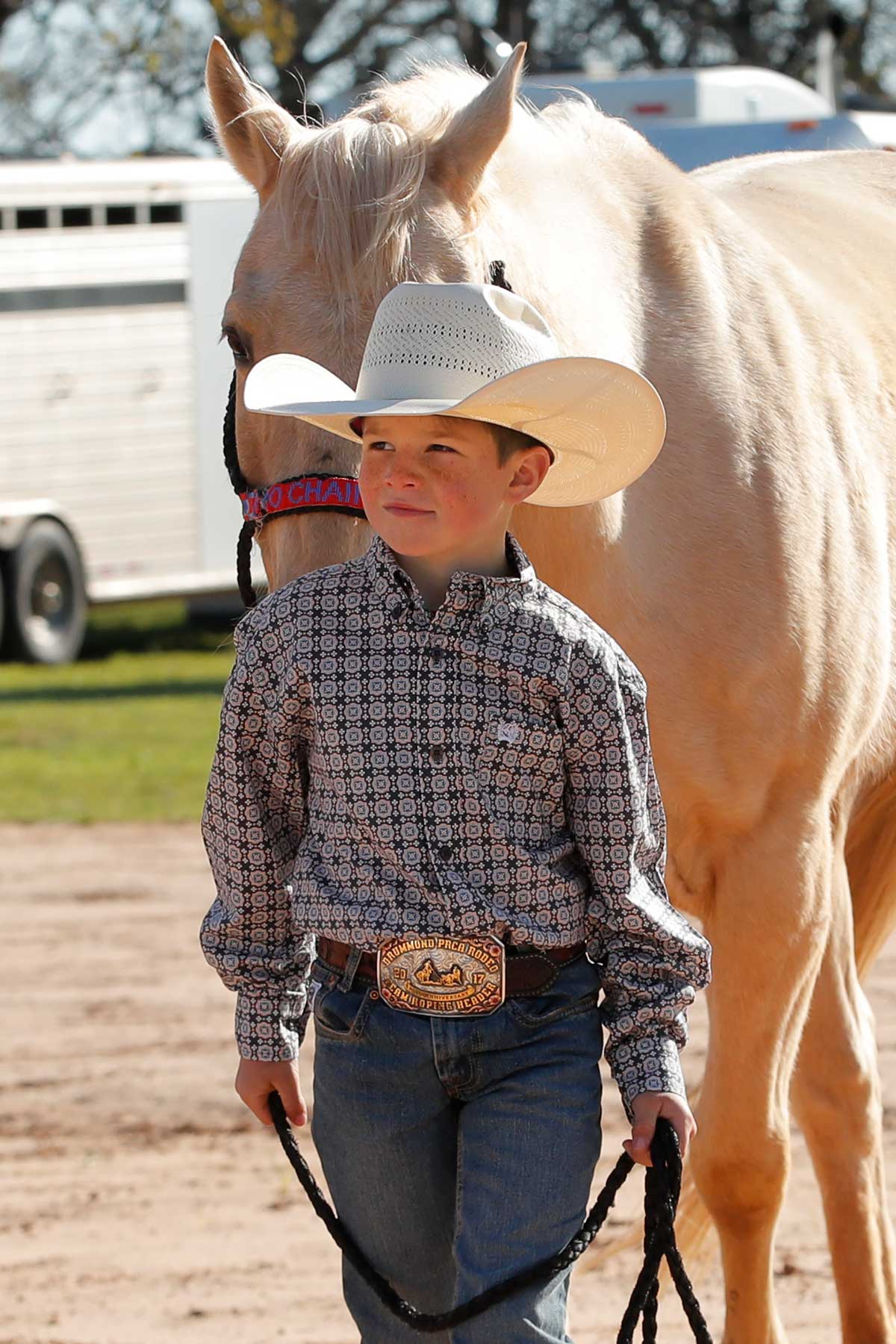 Boy standing outside wearing a gray long sleeve button down shirt, jeans, a belt, and a cowboy hat, leading a horse