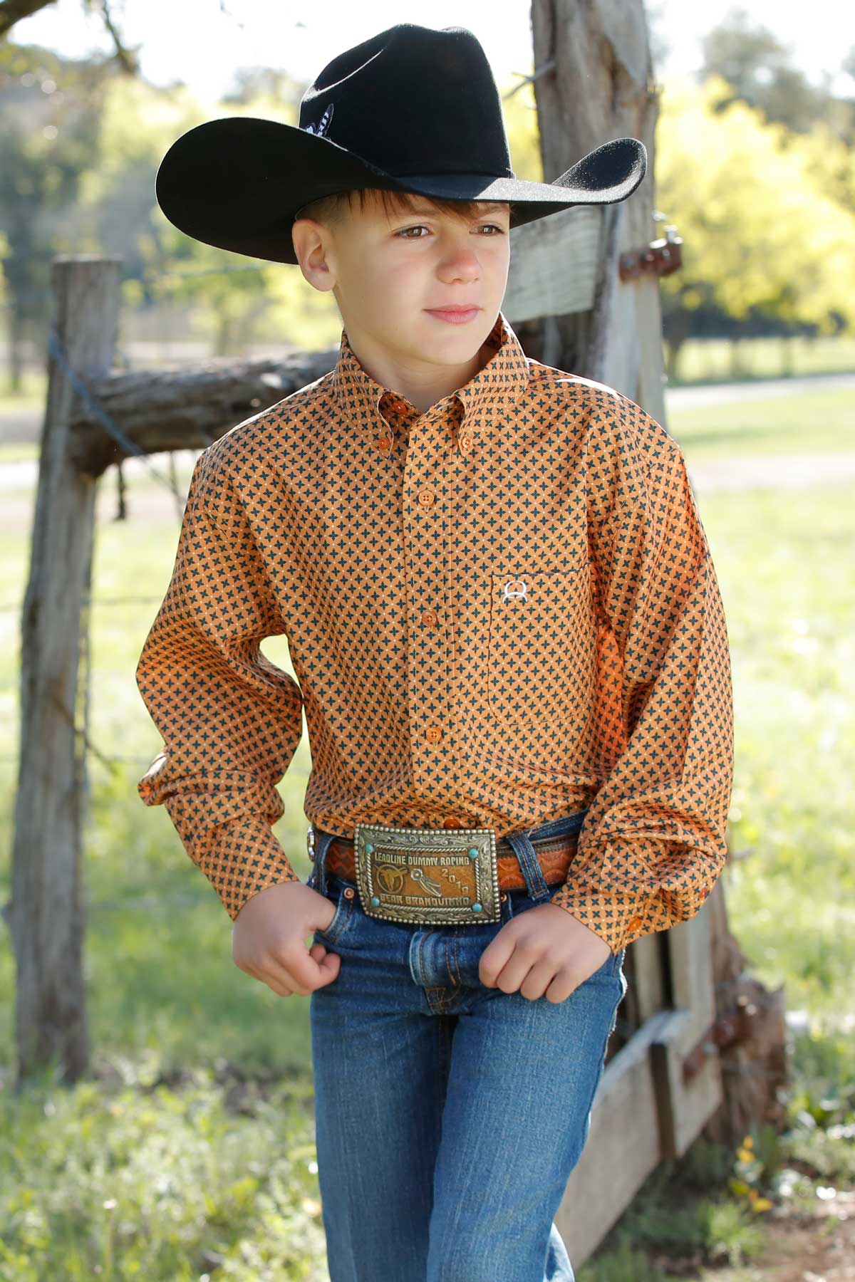 Boy standing outside in front of a fence wearing a orange long sleeve button-down with jeans, a belt and a cowboy hat