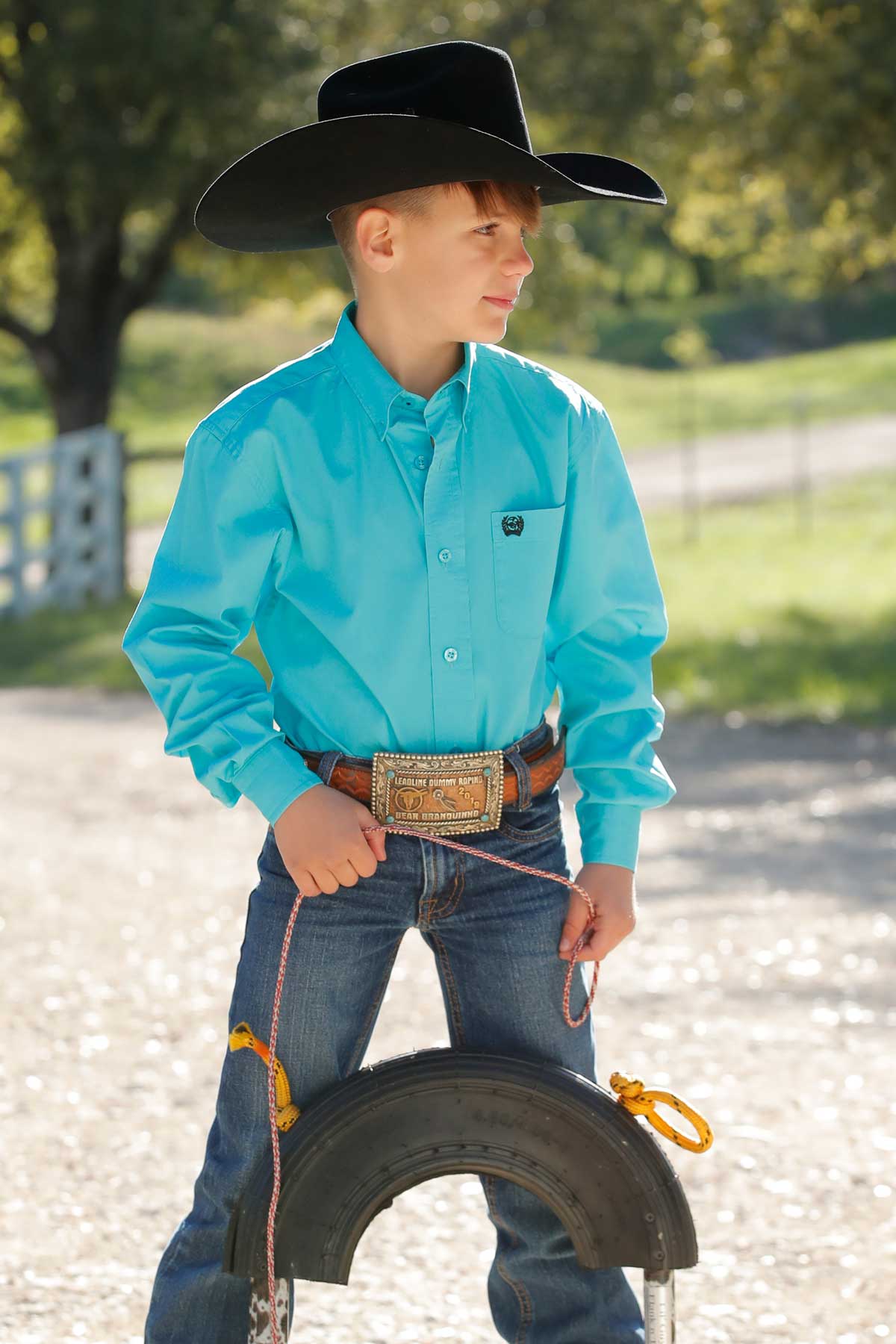 Boy standing outside wearing a teal long sleeve button-down shirt with jeans, a belt, and a cowboy hat, holding a rope