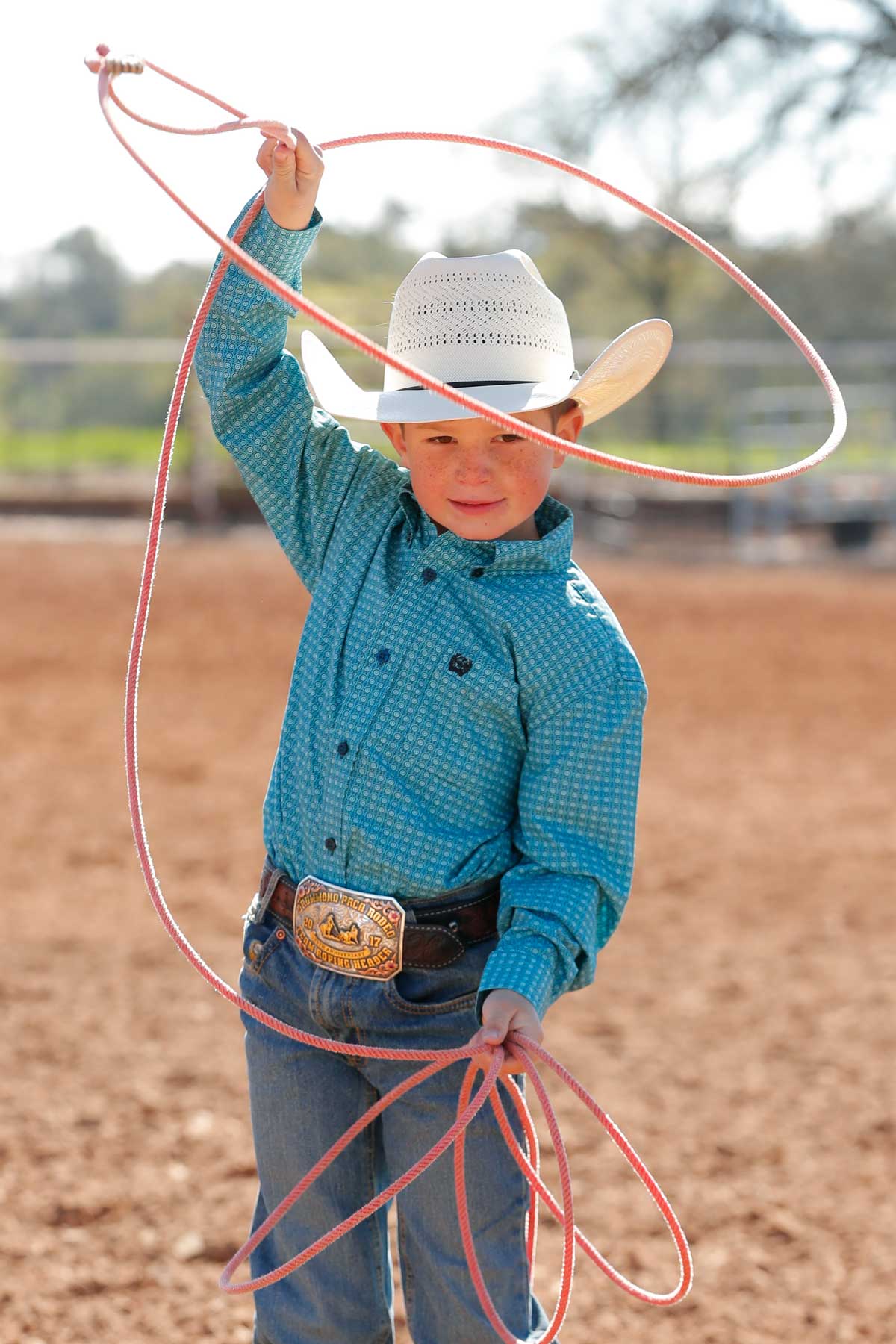 Boy standing outside in an arena wearing a teal long sleeve button down shirt, jeans, a belt, and a cowboy hat holding some rope
