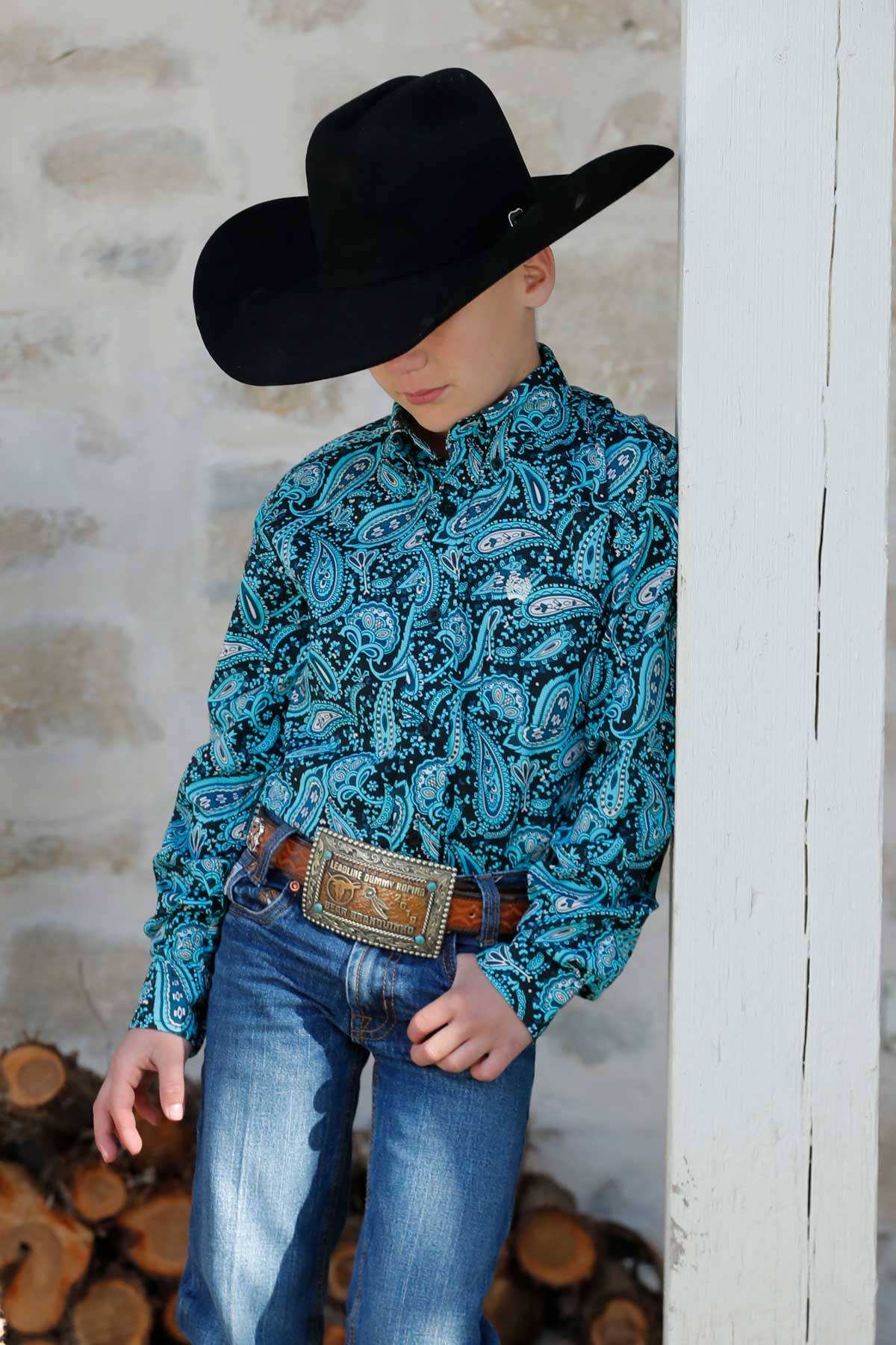 Boy leaning against a wood post wearing a teal and black long sleeve button-down, jeans, a belt, and a cowboy hat