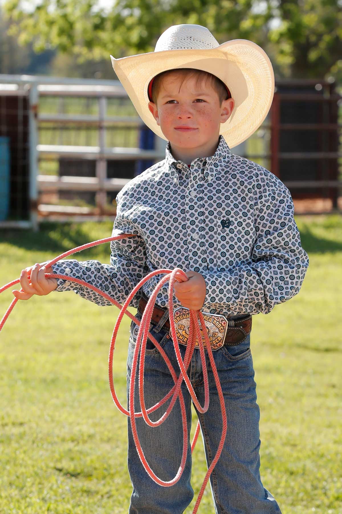 Boy standing outside in front of a fence wearing a long sleeve button down shirt, jeans, a belt, and a cowboy hat holding some rope