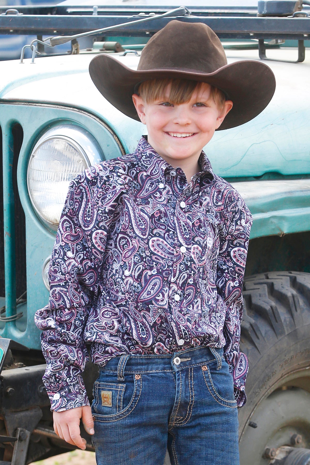 Boy standing outside in front of a car wearing a purple long sleeve button down shirt, jeans, a belt, and a cowboy hat