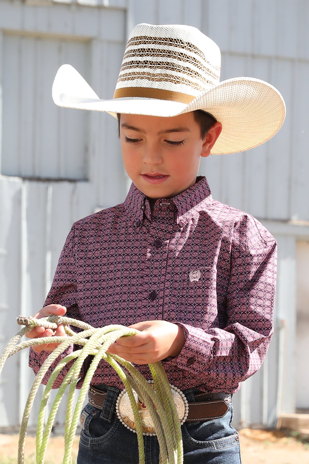 Boy standing outside in front of a barn wearing a purple long sleeve button down shirt, jeans, a belt, and a cowboy hat holding some rope
